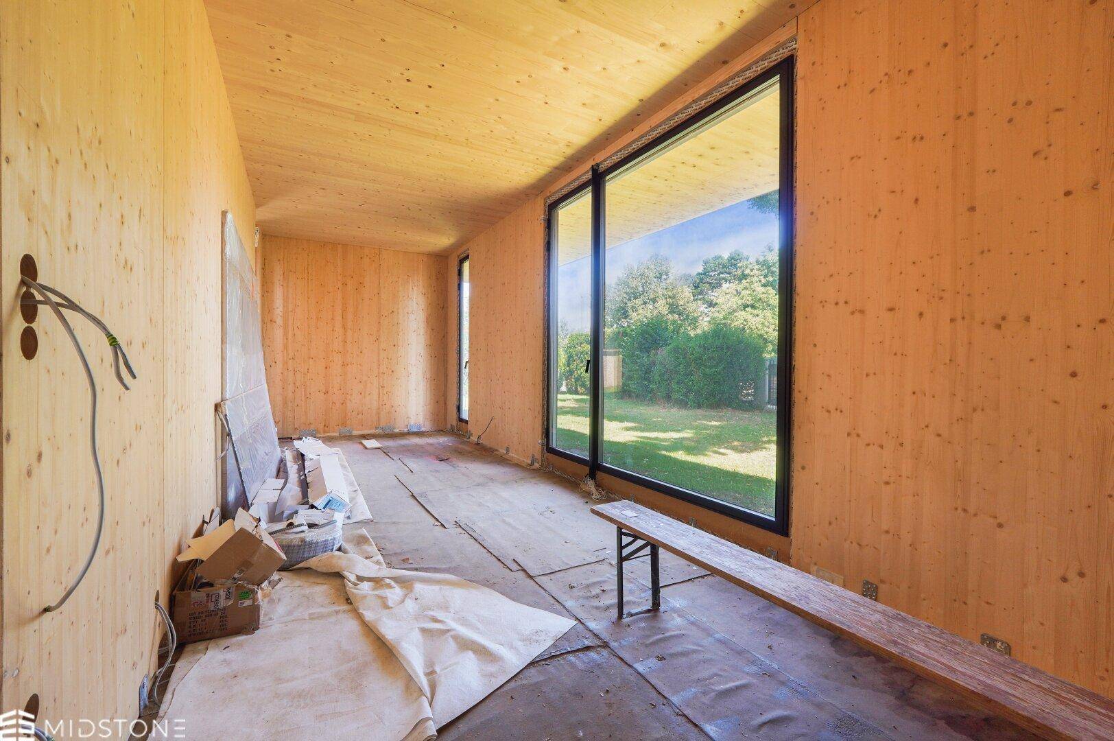 Innenraum im Rohbau mit Holzverkleidung, großen Schiebefenstern und Blick auf den Garten.