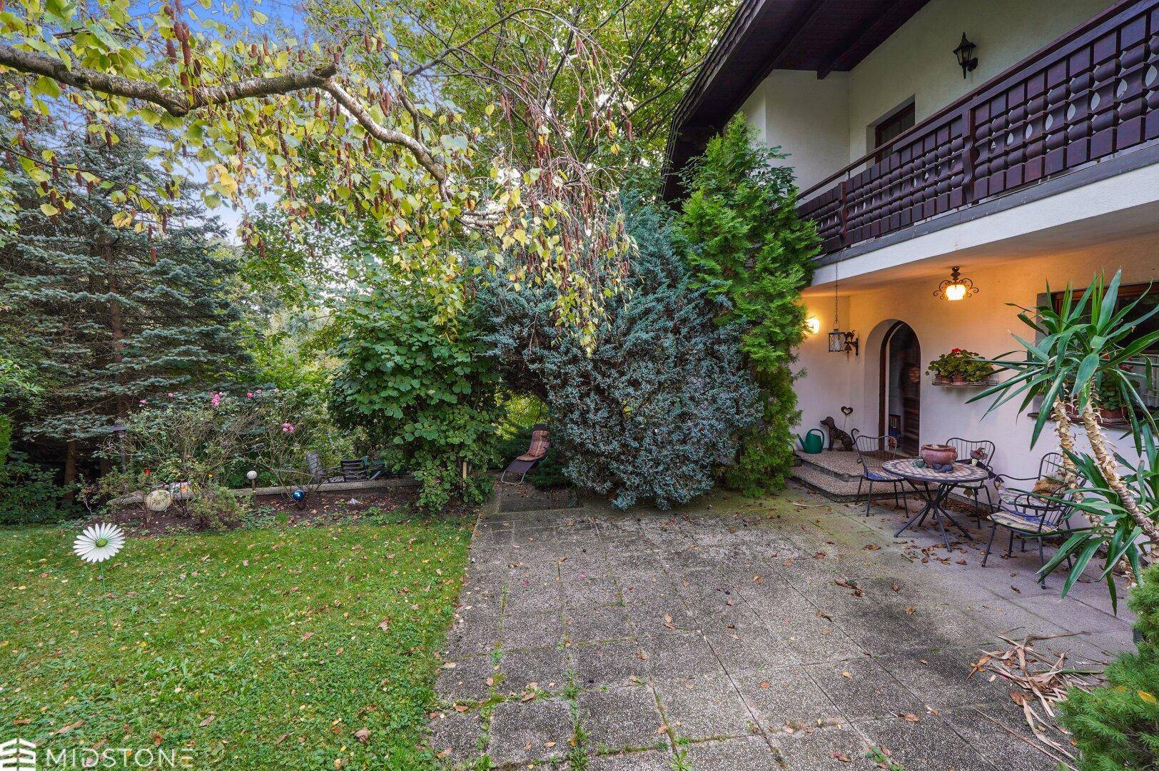 Großer Gartenbereich mit Rasenfläche und gepflasterter Terrasse vor dem Haus.