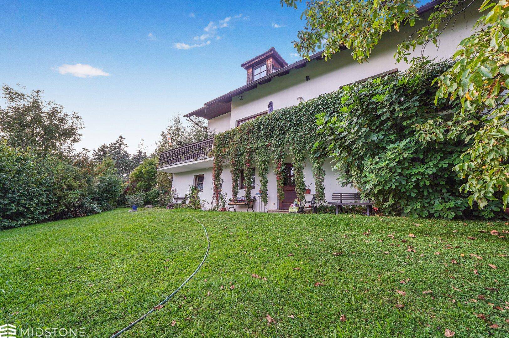 Hausfassade mit Kletterpflanzen und weitem Garten, unter blauem Himmel.