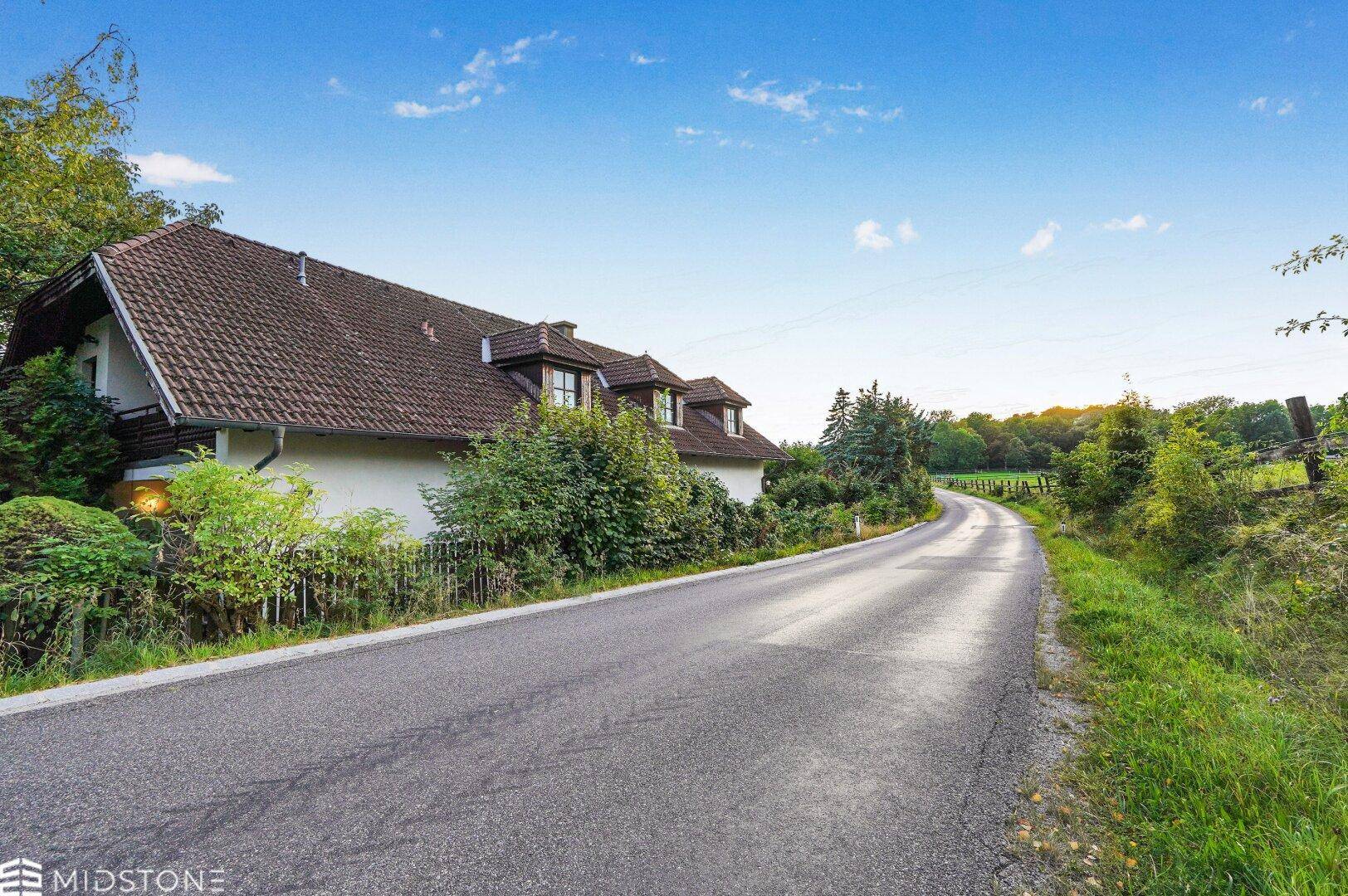 Landstraße führt am Haus vorbei, umgeben von grüner Landschaft und Bäumen.