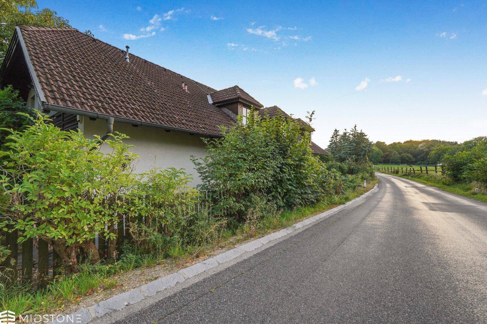 Haus an einer Landstraße, umgeben von Natur und Bäumen unter blauem Himmel.