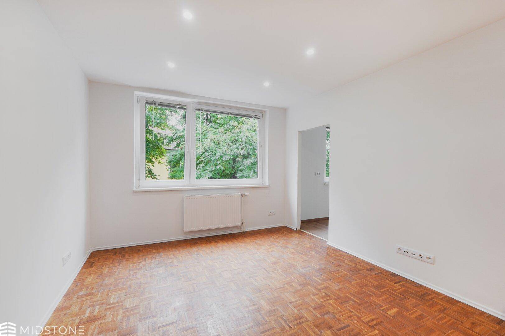 Geräumiges Zimmer mit hellem Parkett und großem Fenster mit Blick ins Grüne.