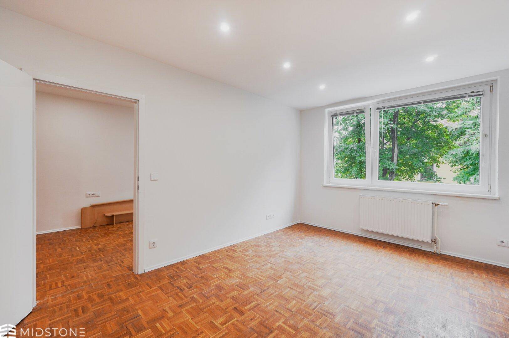 Großzügiges Zimmer mit Parkettboden und Fensterfront, die viel Tageslicht hereinlässt.