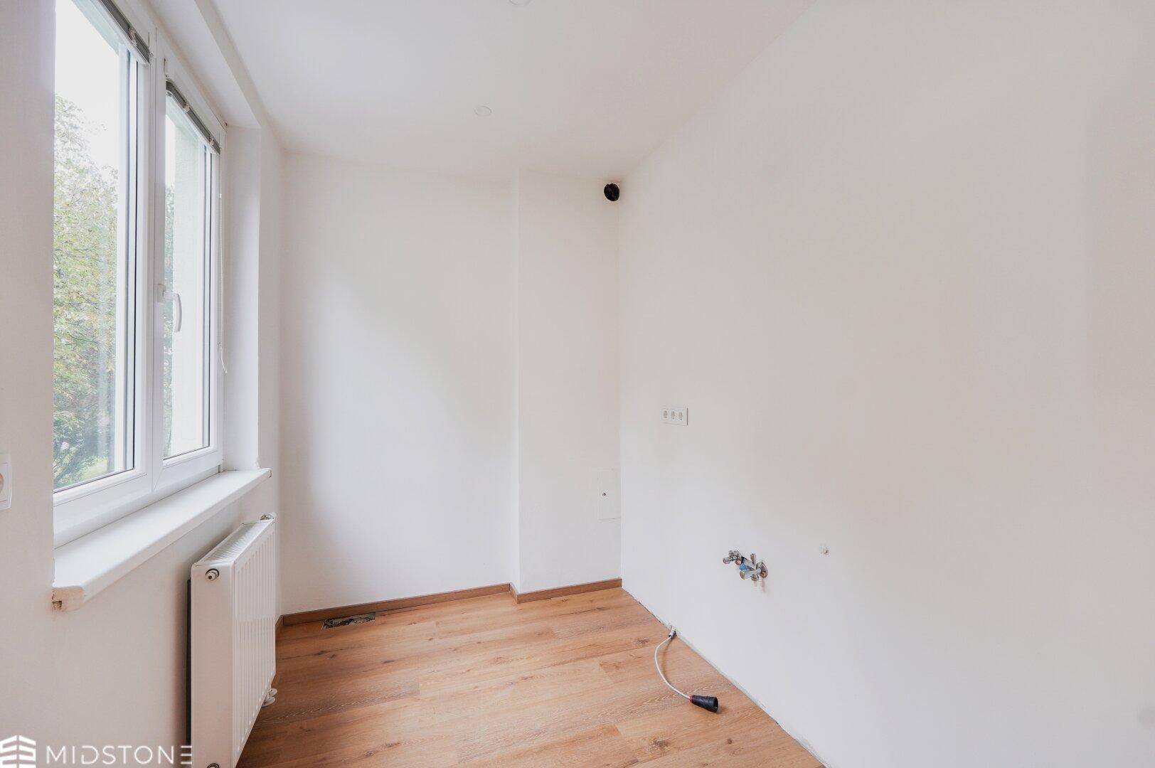 Leere Küche mit vorbereiteten Anschlüssen und hellem Holzboden, bereit für individuelle Gestaltung.