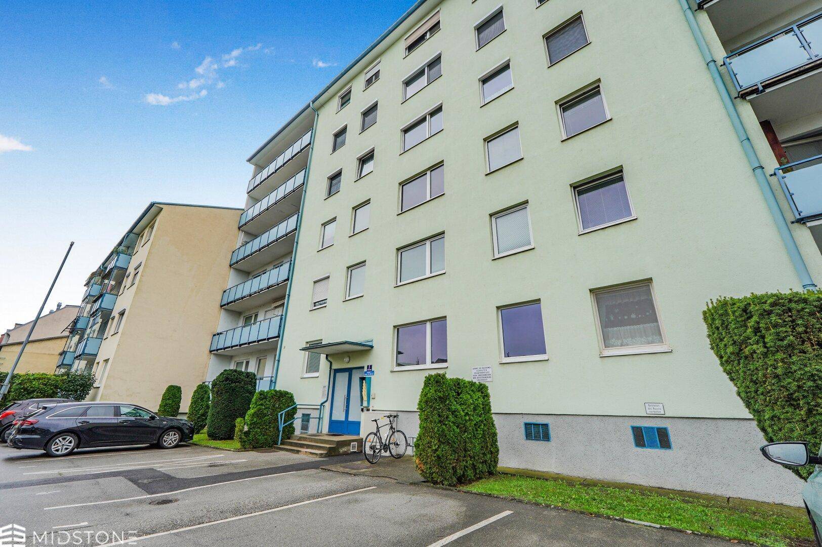 Wohngebäude mit mehreren Etagen, Balkonen und Parkmöglichkeiten vor dem Eingang.