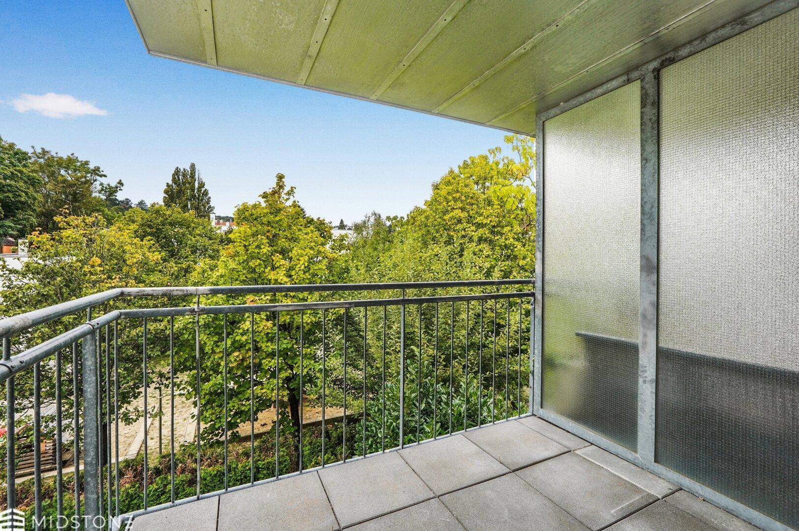 Heller Balkon mit Fliesenboden und Blick ins Grüne, umgeben von Bäumen.