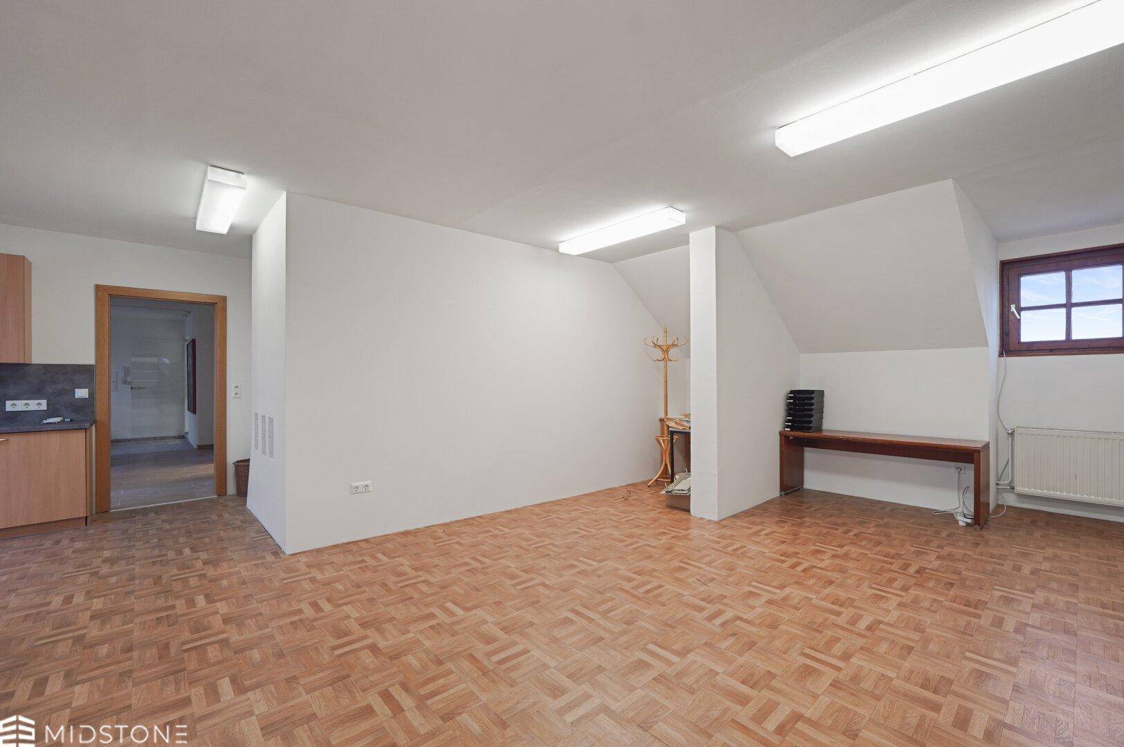 Offener Bürobereich mit Parkettboden, Dachschrägen und einem kleinen Fenster, mit Blick auf eine Küchenzeile.
