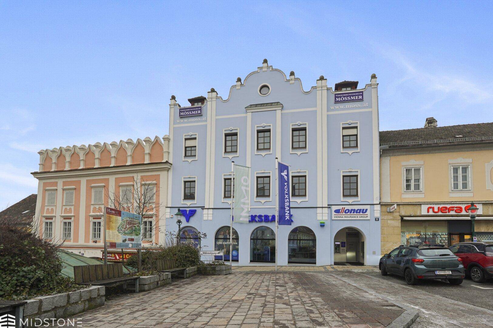 Historisches Gebäude mit blauer Fassade und Geschäften im Erdgeschoss, gelegen an einem öffentlichen Platz.