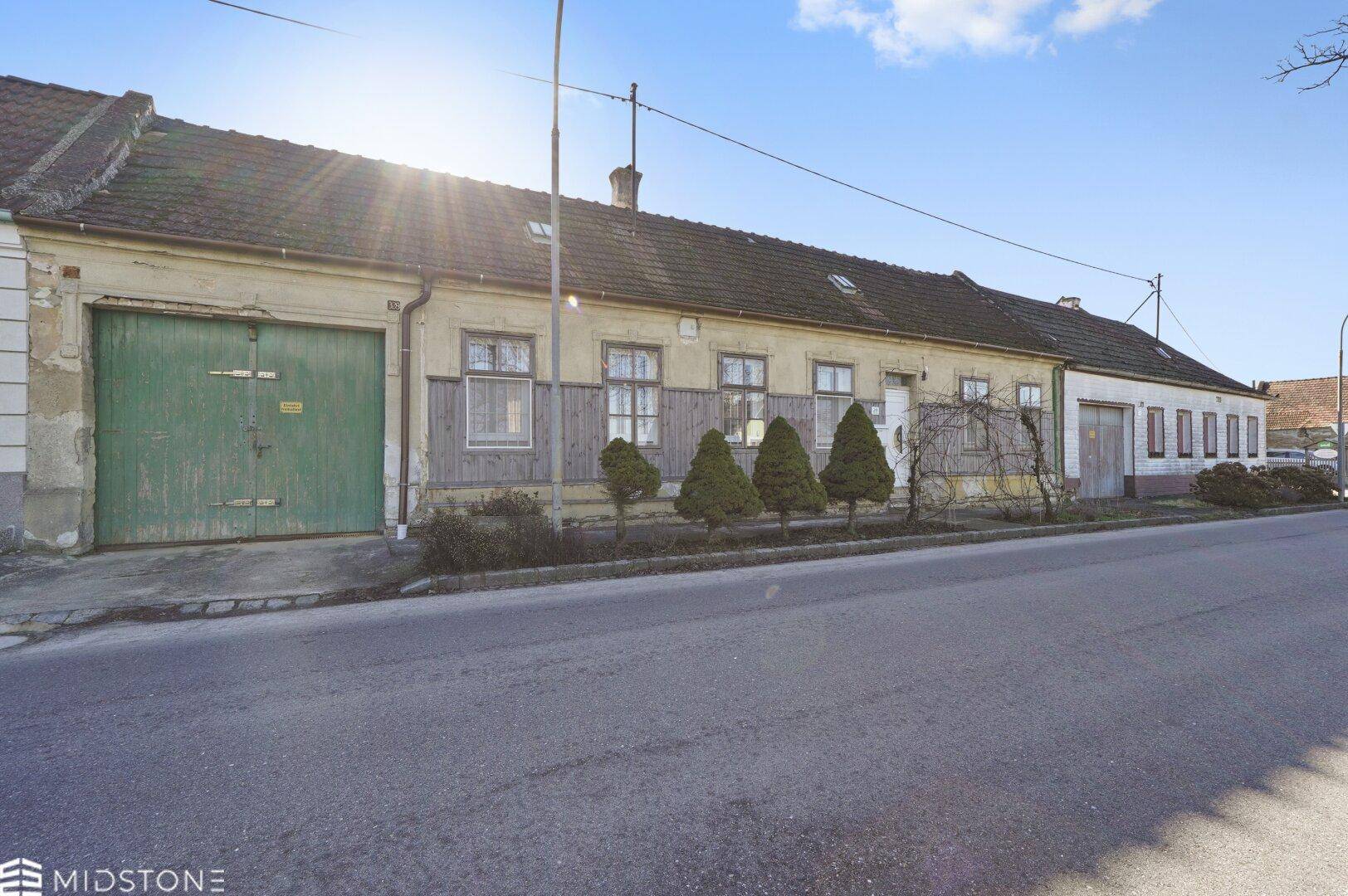 Langes, älteres Wohnhaus mit gelblicher Fassade und grünen Garagentoren an einer ruhigen Straße.