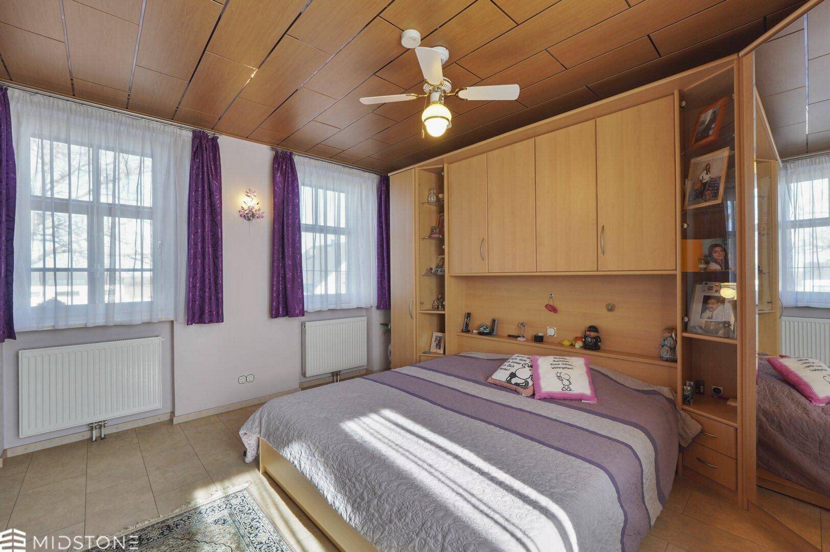 Helles Schlafzimmer mit zwei Fenstern, Deckenventilator und Einbauschränken.