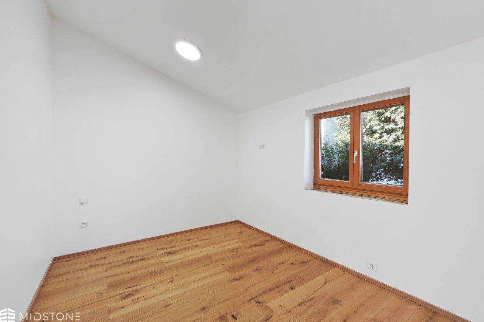 Leeres Zimmer mit Holzboden, weißen Wänden und einem Fenster mit Blick ins Grüne.