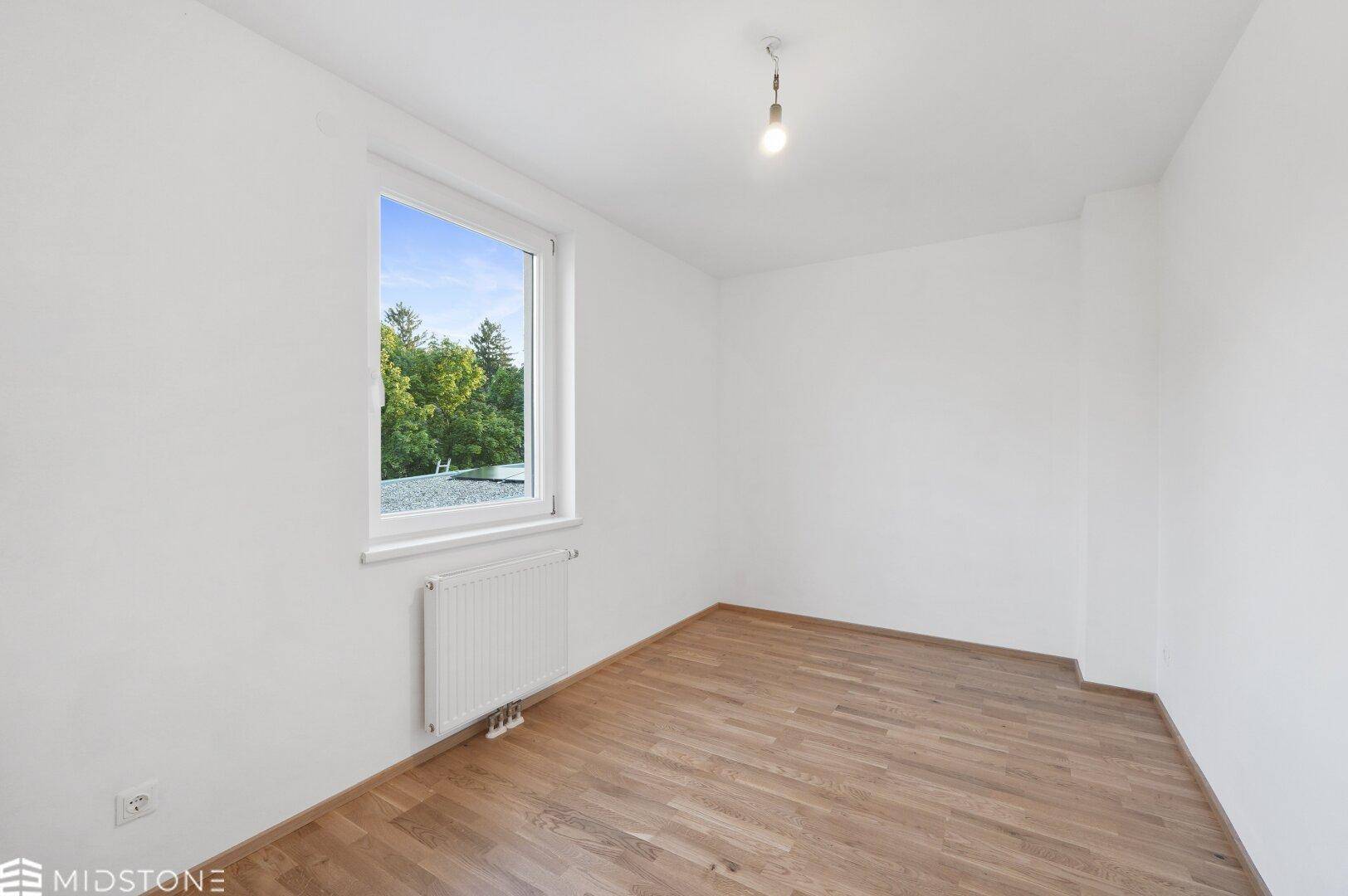 Helles Schlafzimmer mit Parkettboden und einem großen Fenster mit Blick ins Grüne.