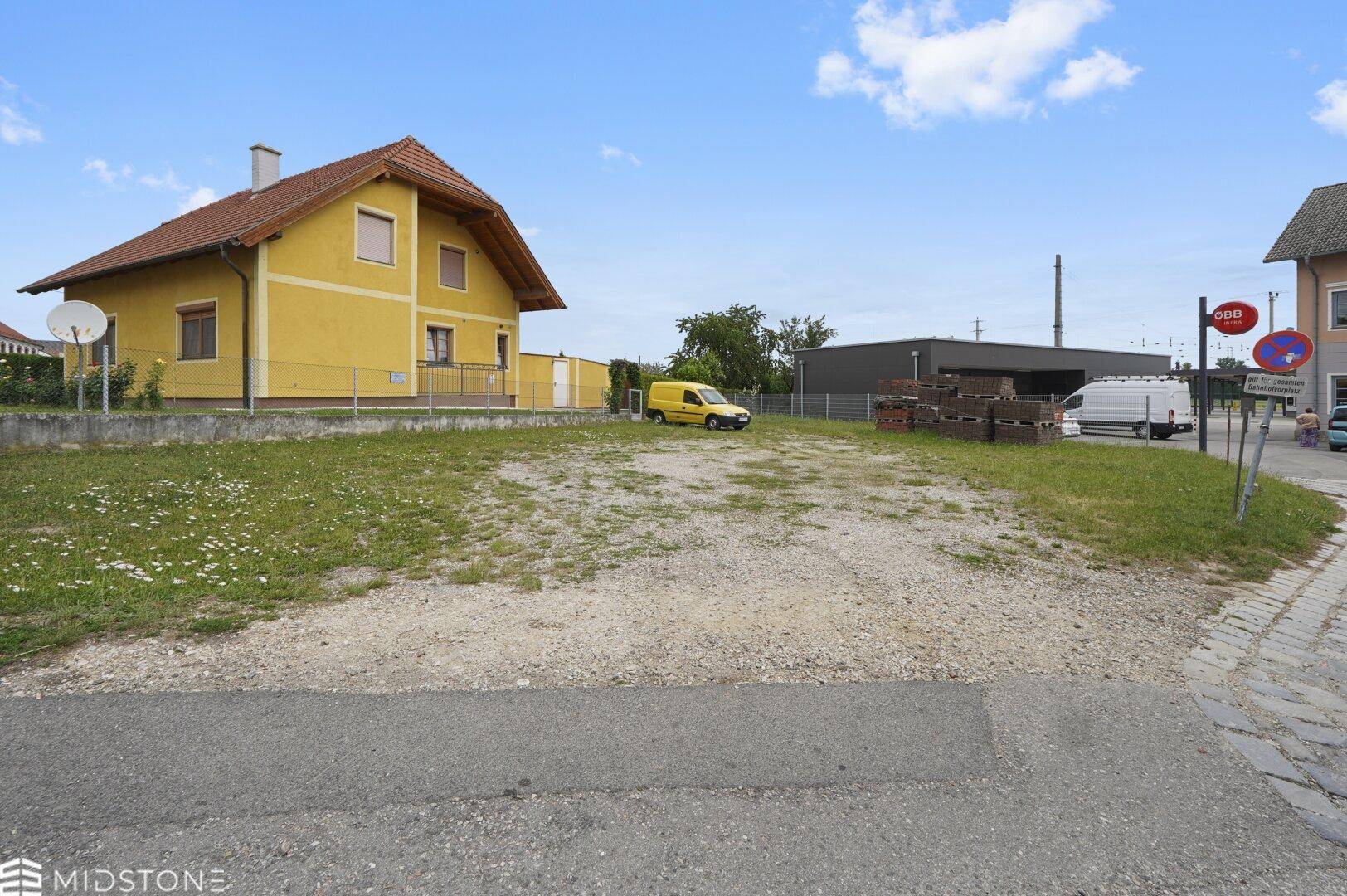 Blick auf ein unbebautes Grundstück mit einem gelben Haus im Hintergrund und weiteren Gebäuden in der Umgebung.
