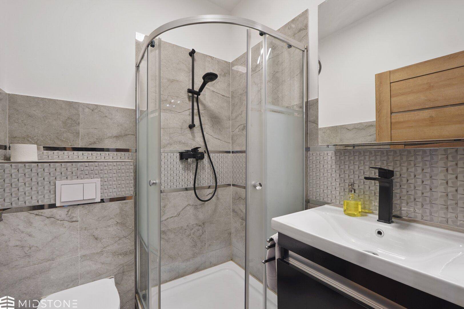 Modernes Badezimmer mit Duschkabine, Waschbecken und grauen Fliesen, ausgestattet mit schwarzen Armaturen.
