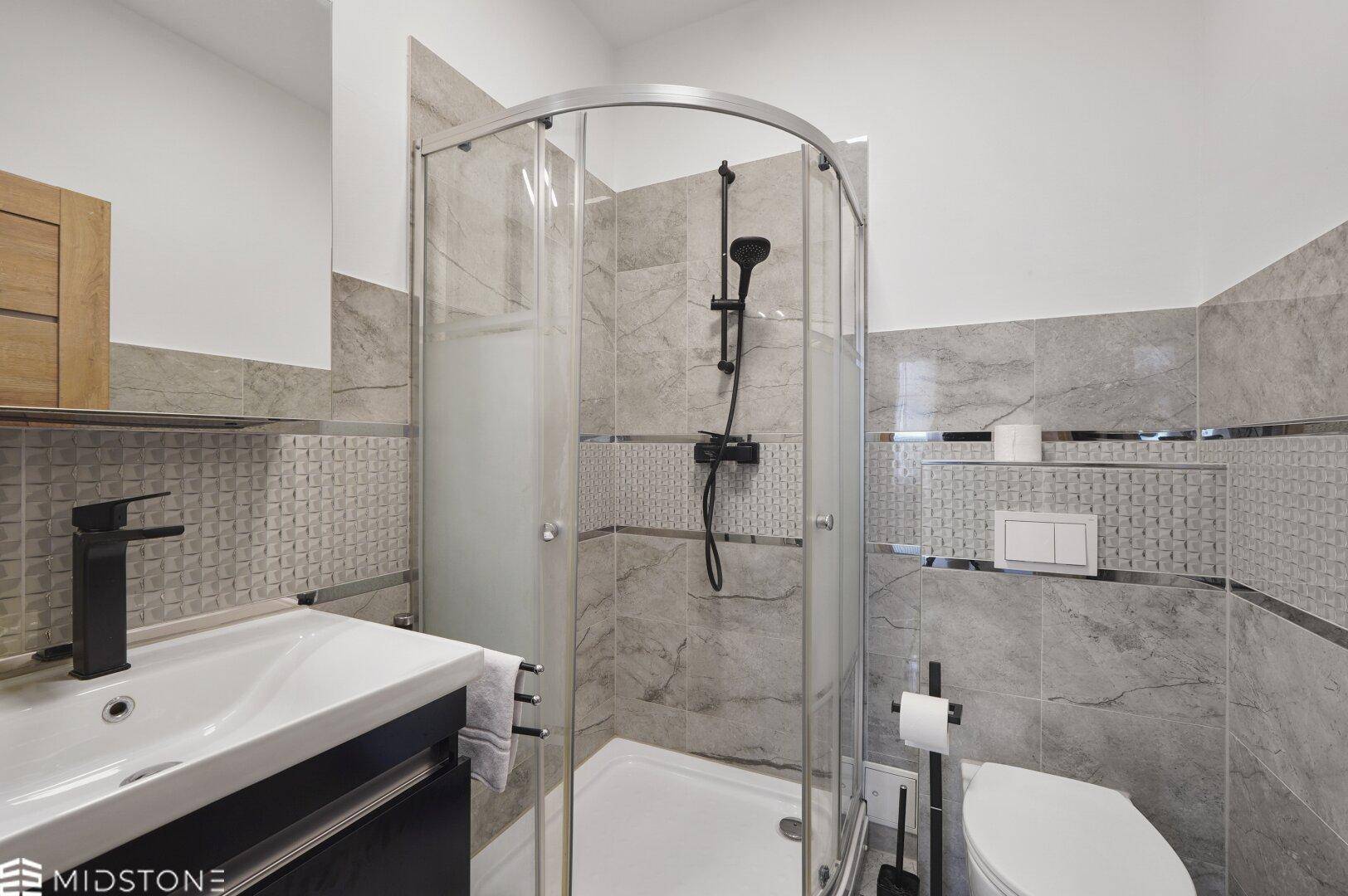 Modernes Badezimmer mit Duschkabine, Waschbecken und grauen Fliesen, mit dunklen Armaturen.