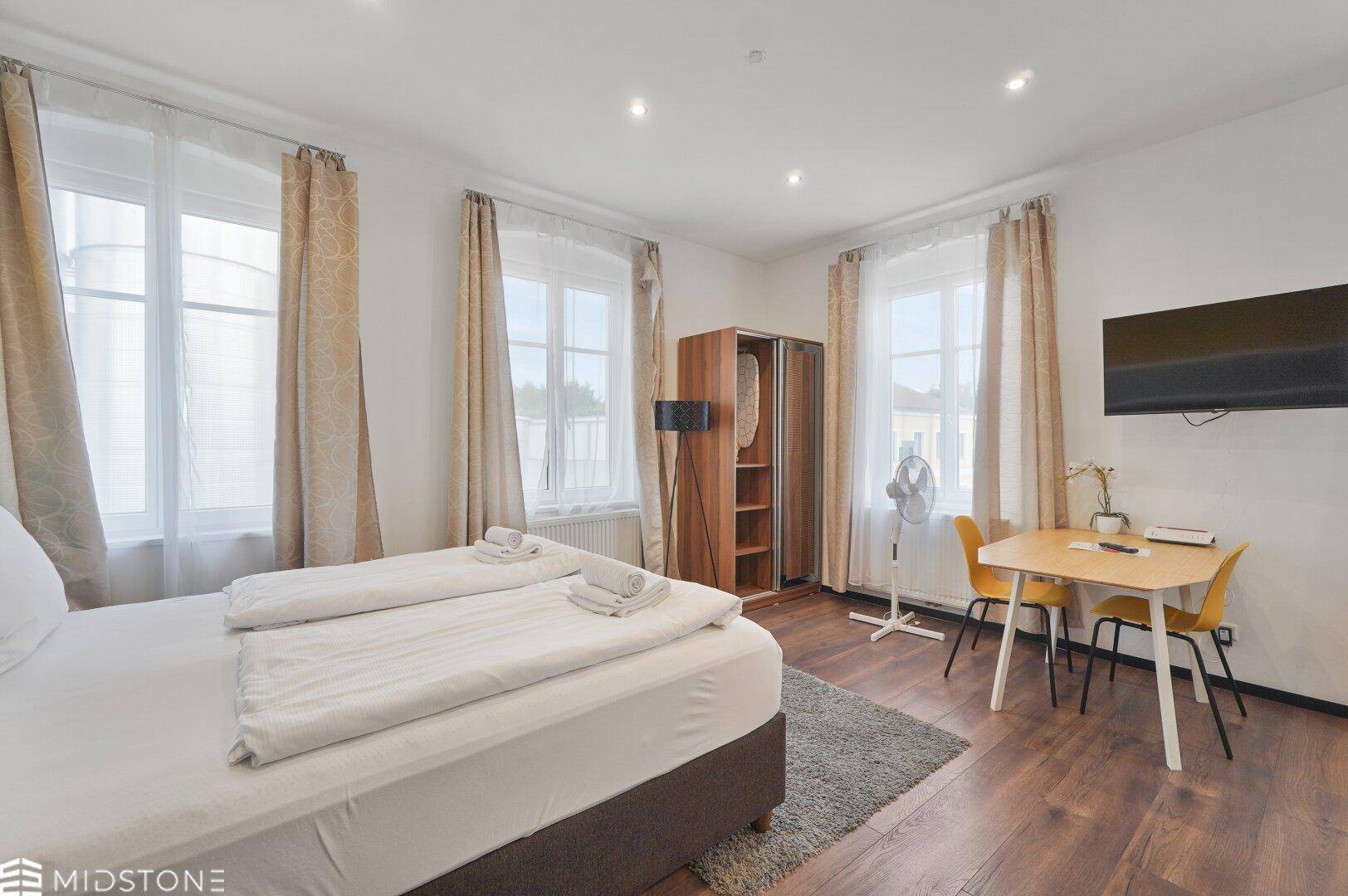 Helles Schlafzimmer mit Doppelbett, Kleiderschrank und kleinem Esstisch, ideal für Kurzaufenthalte.