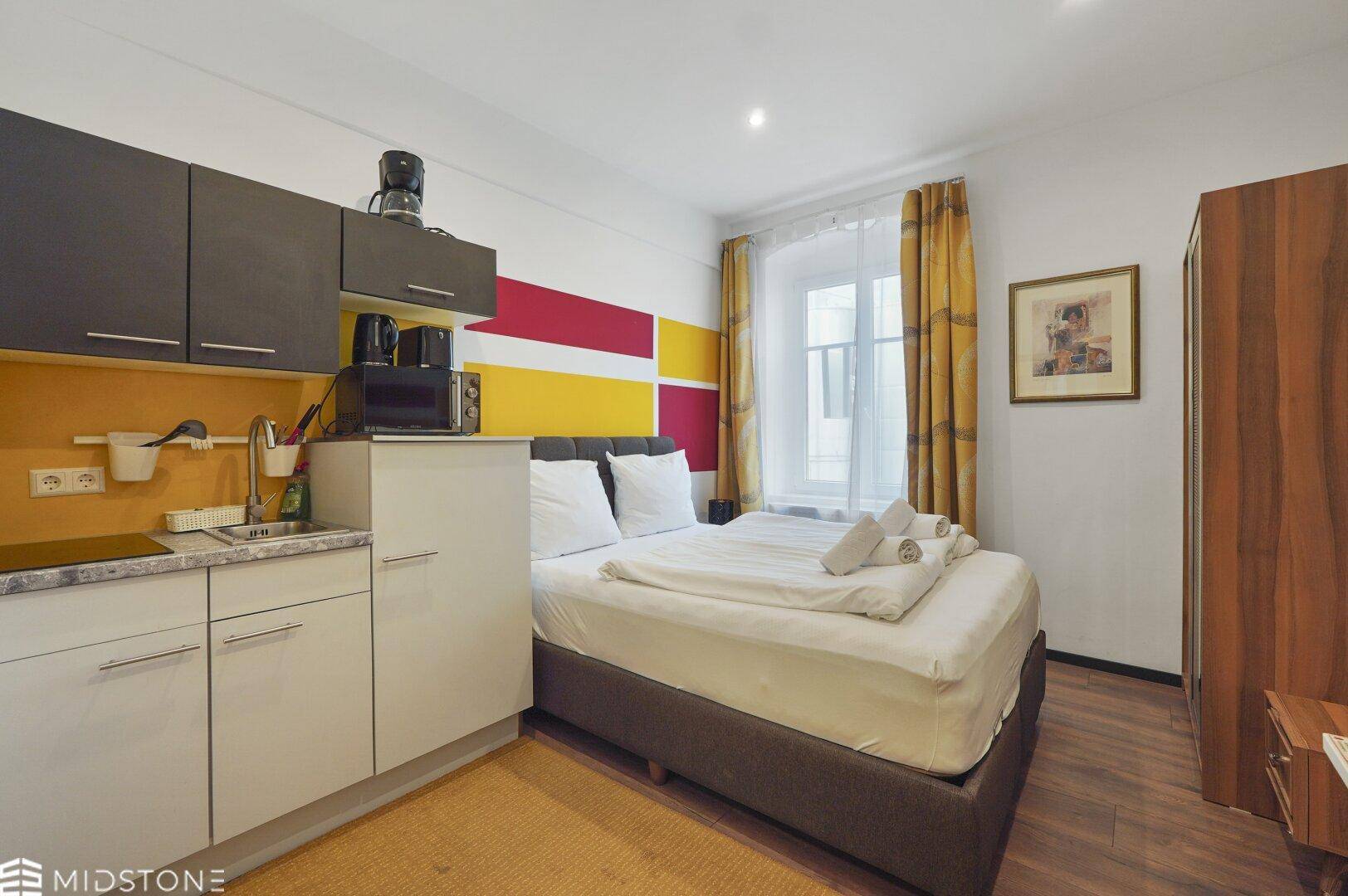 Kompaktes Studio-Apartment mit Küchenzeile und Schlafbereich, farbenfroh gestaltet mit roten Akzenten.
