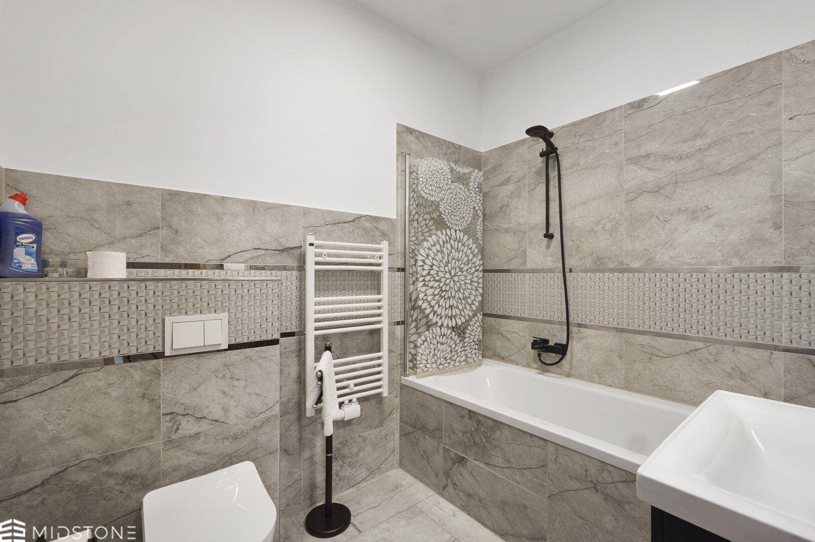 Modernes Badezimmer mit Badewanne, grauen Fliesen und Handtuchheizkörper, sauber und funktional gestaltet.