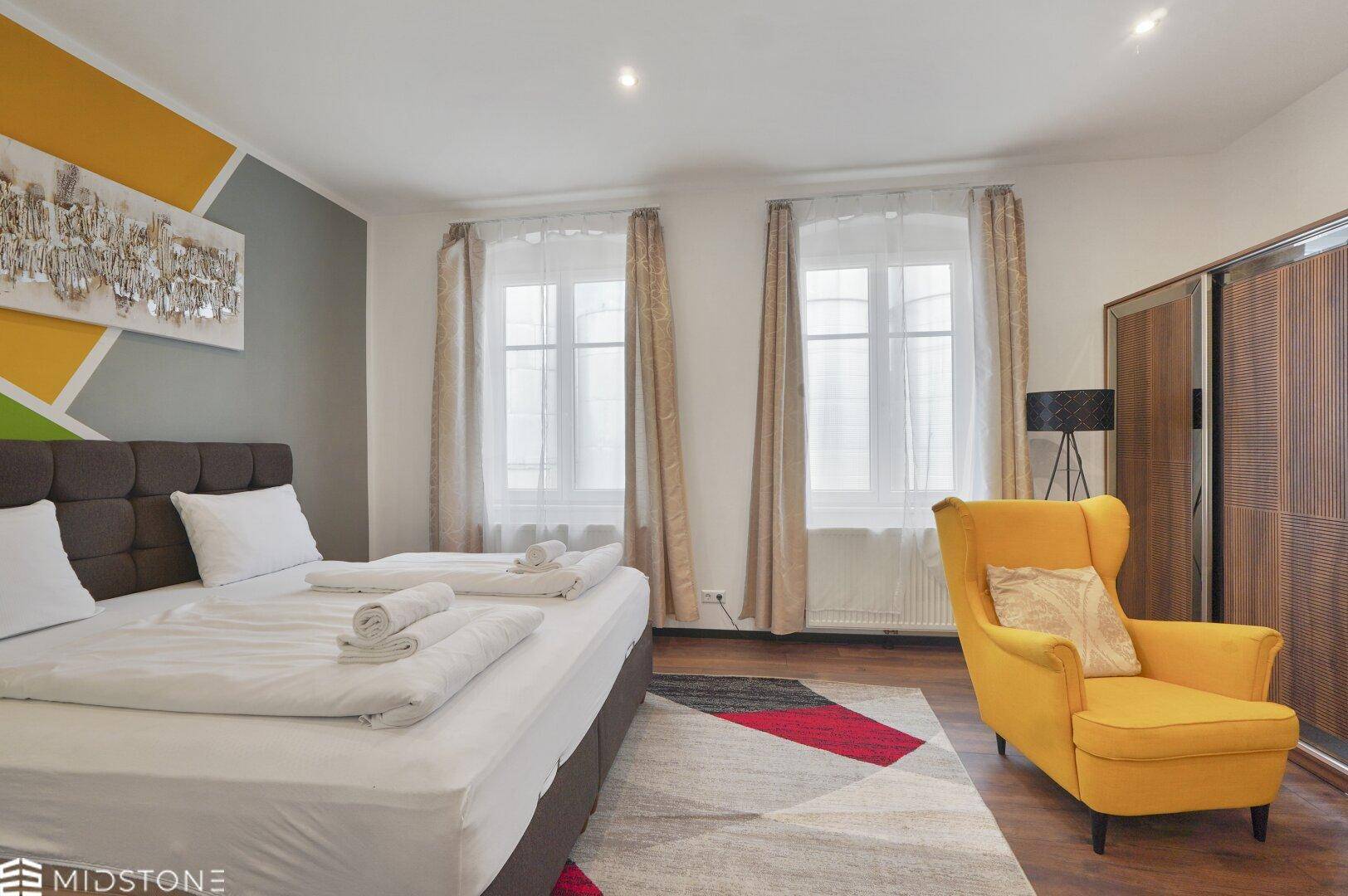 Helles Schlafzimmer mit Doppelbett, gelbem Sessel und großem Kleiderschrank in modernem Stil.