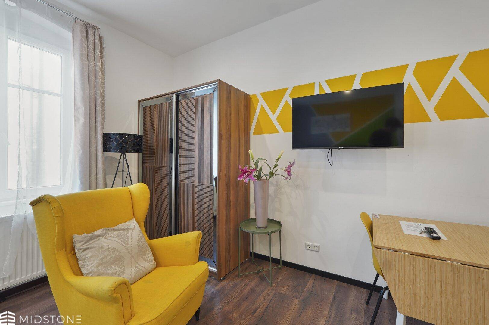 Schlafzimmer mit Wandfernseher, gelbem Sessel und modernem Kleiderschrank, akzentuiert durch gelbe Muster.