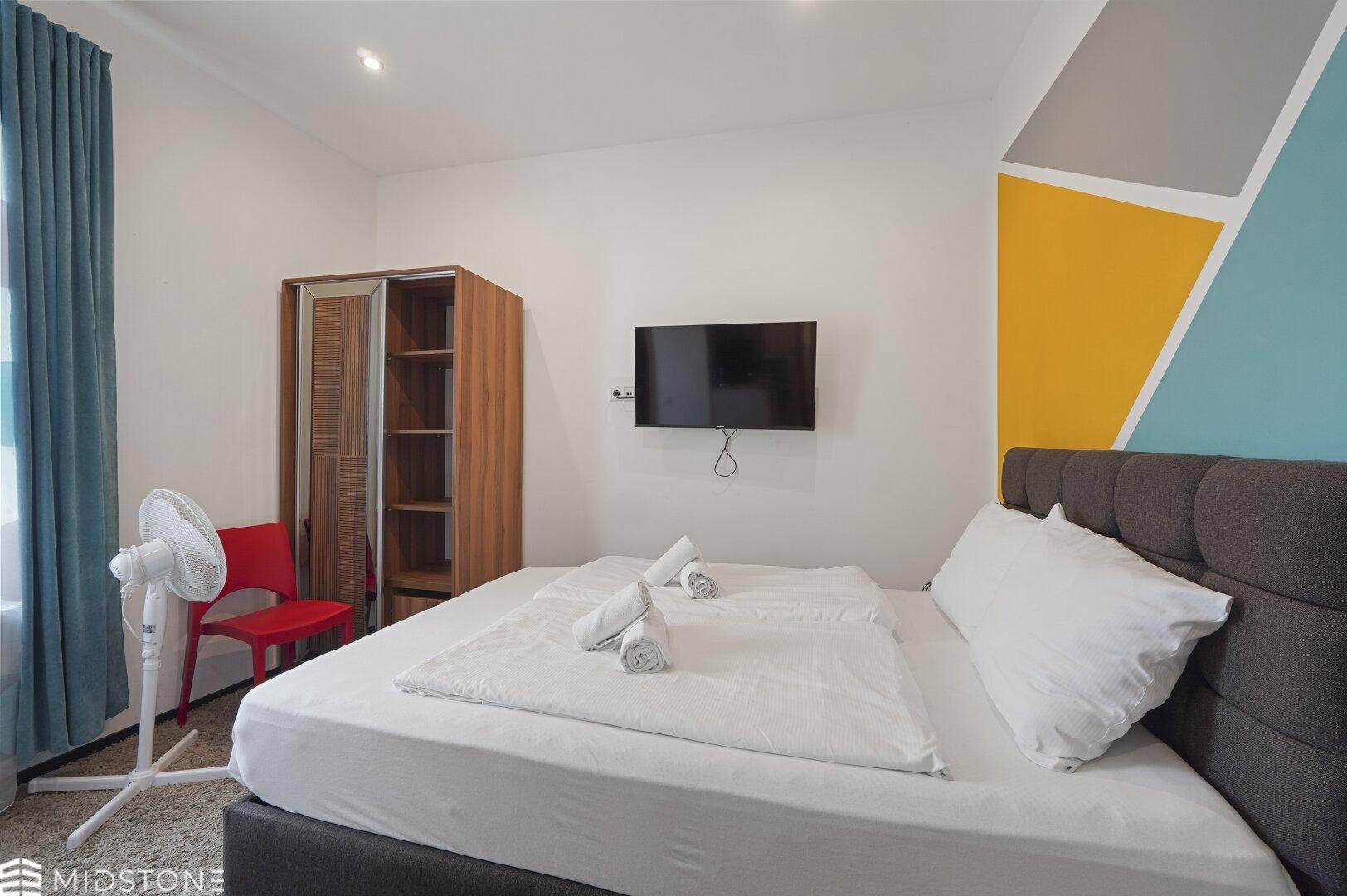 Schlafzimmer mit Doppelbett, Wandfernseher und farbiger Akzentwand, modern und einladend gestaltet.