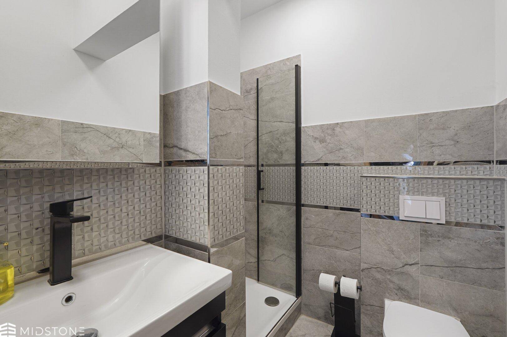 Modernes Badezimmer mit Duschbereich, Waschbecken und grauen Fliesen, mit schwarzen Armaturen.