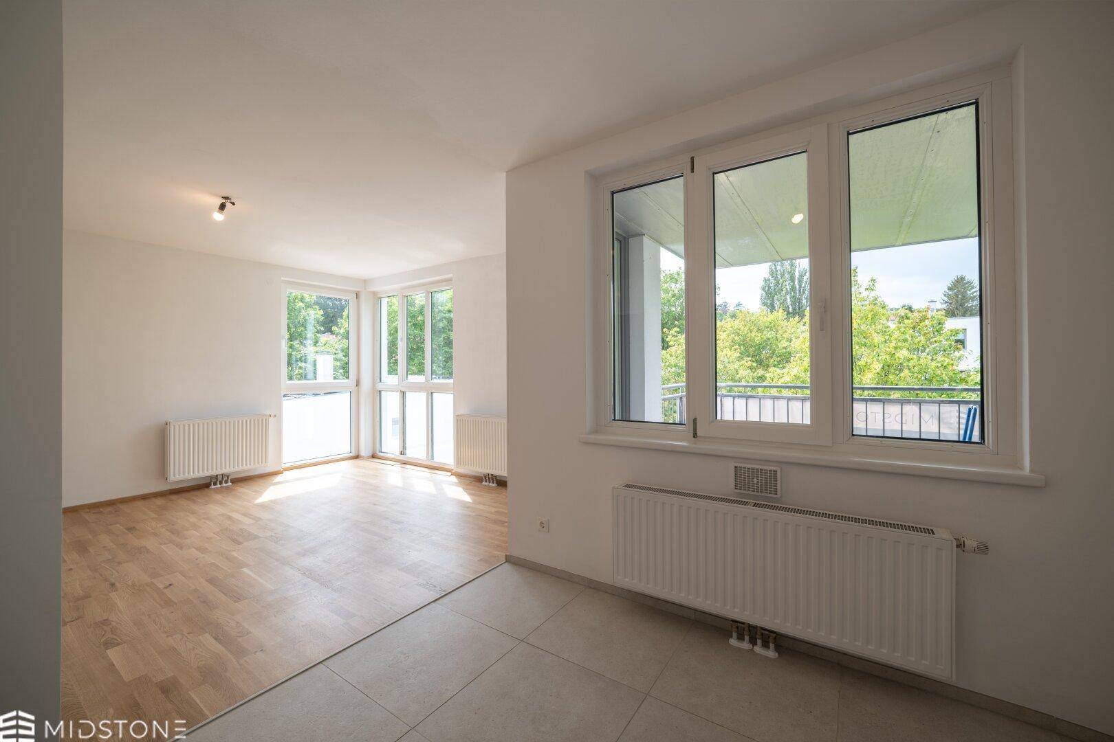 Offener Wohnbereich mit Parkett- und Fliesenboden, großen Fenstern und Balkonzugang.