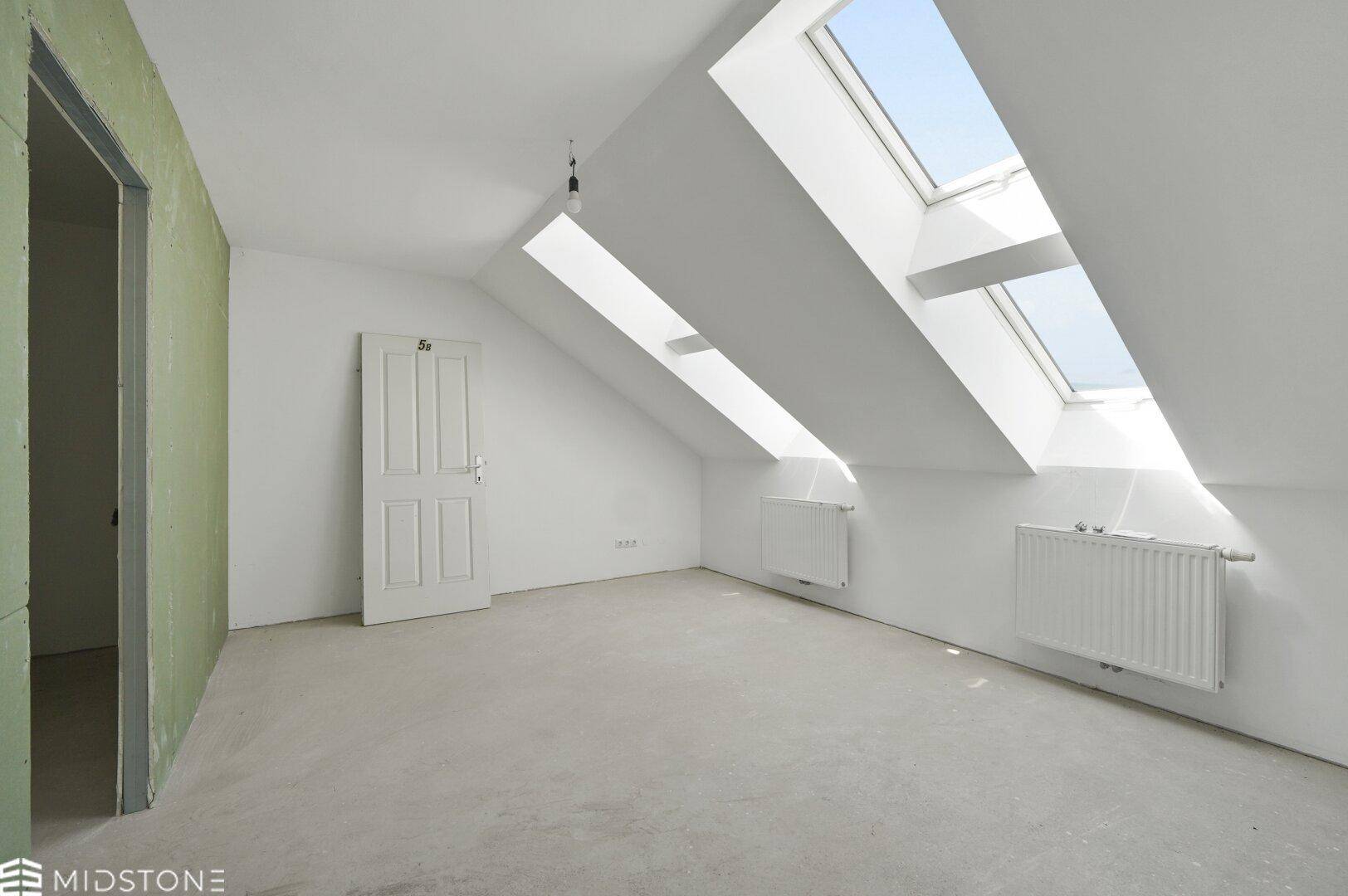 Helles Dachzimmer mit schrägen Wänden, mehreren Dachfenstern und Betonboden.