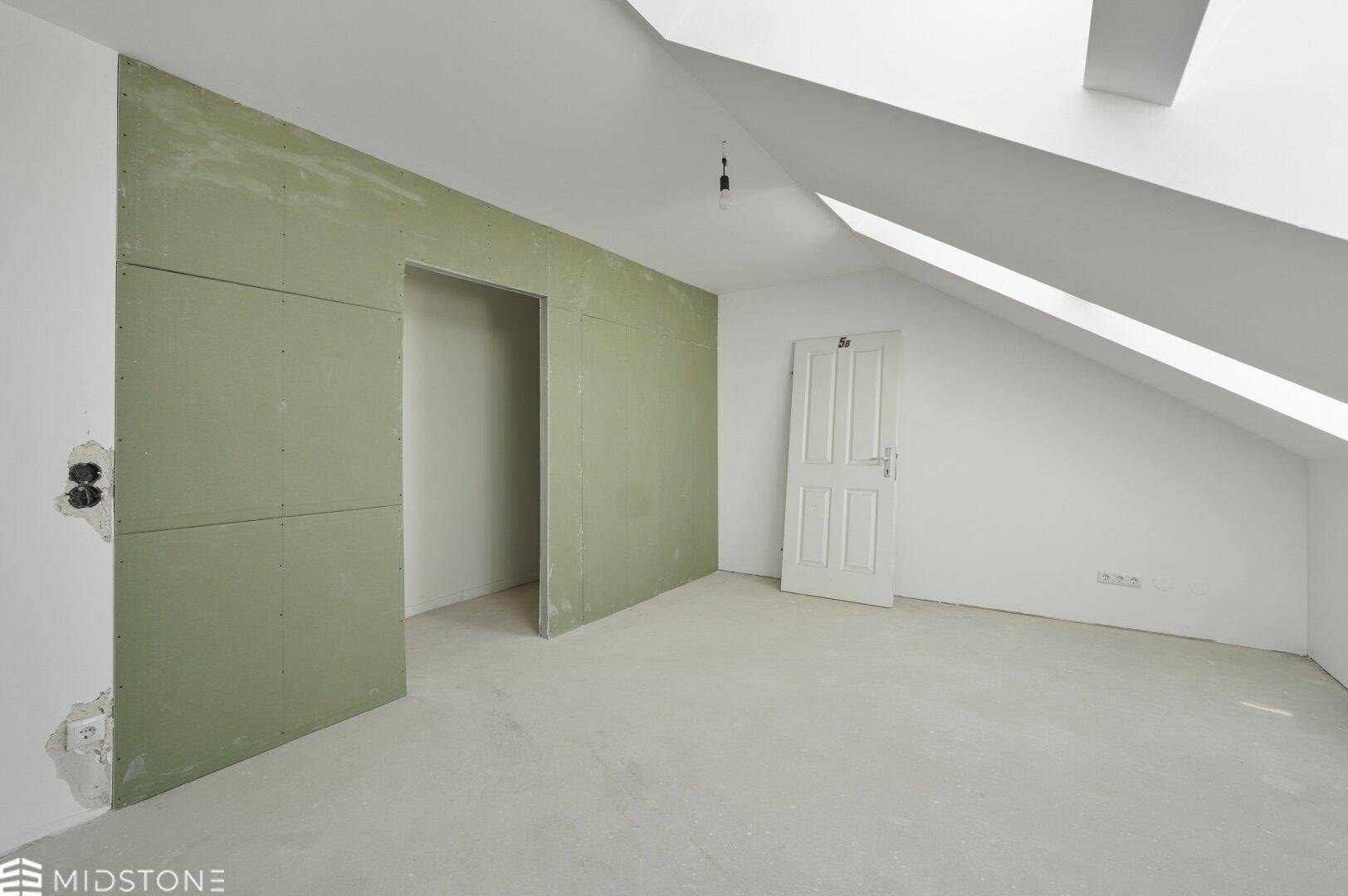 Dachzimmer mit schrägen Wänden, Betonboden und vorbereiteter Wand für Einbauten.