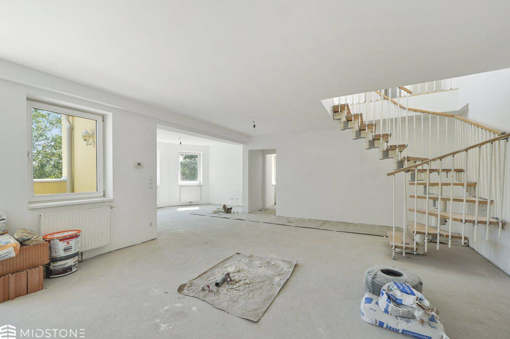 Geräumiger Wohnbereich im Rohzustand mit viel Tageslicht und einer geschwungenen Treppe.