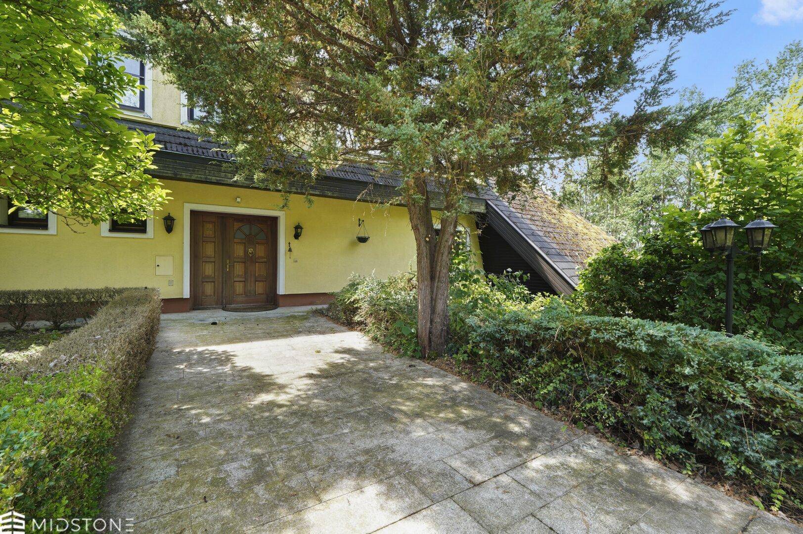 Eingangsbereich des Hauses mit gepflastertem Weg, doppelten Holztüren und gepflegten Hecken.