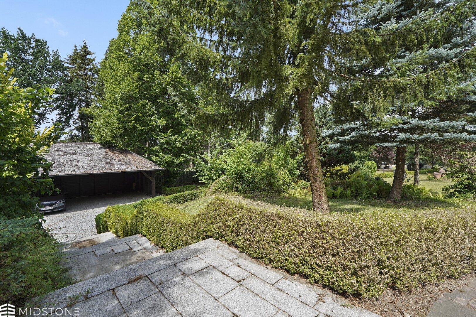 Ansicht des Anwesens mit Carport, hohen Bäumen und gepflegten Hecken.
