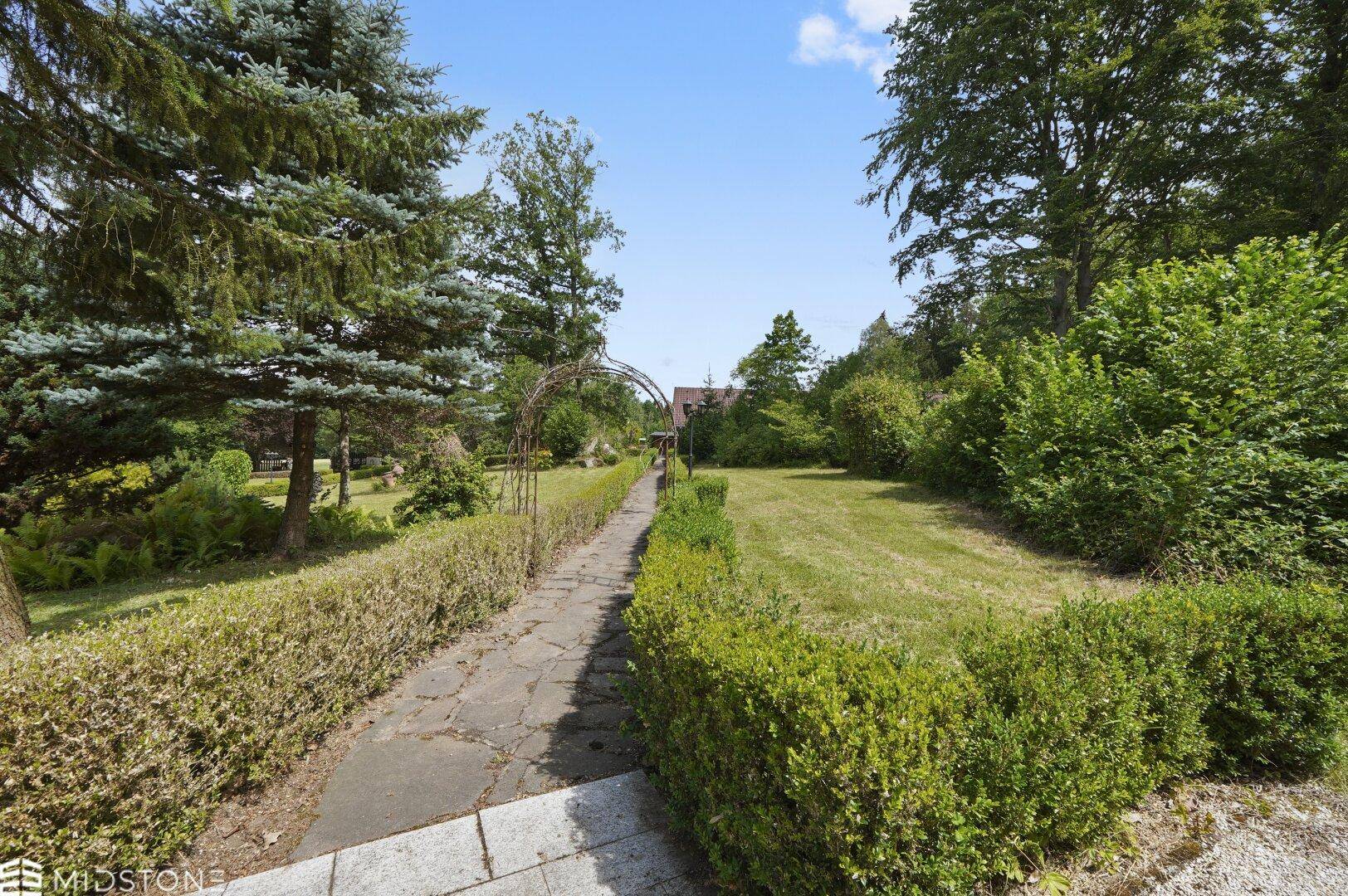 Steinweg führt durch einen gepflegten Garten mit Hecken und einem Torbogen.
