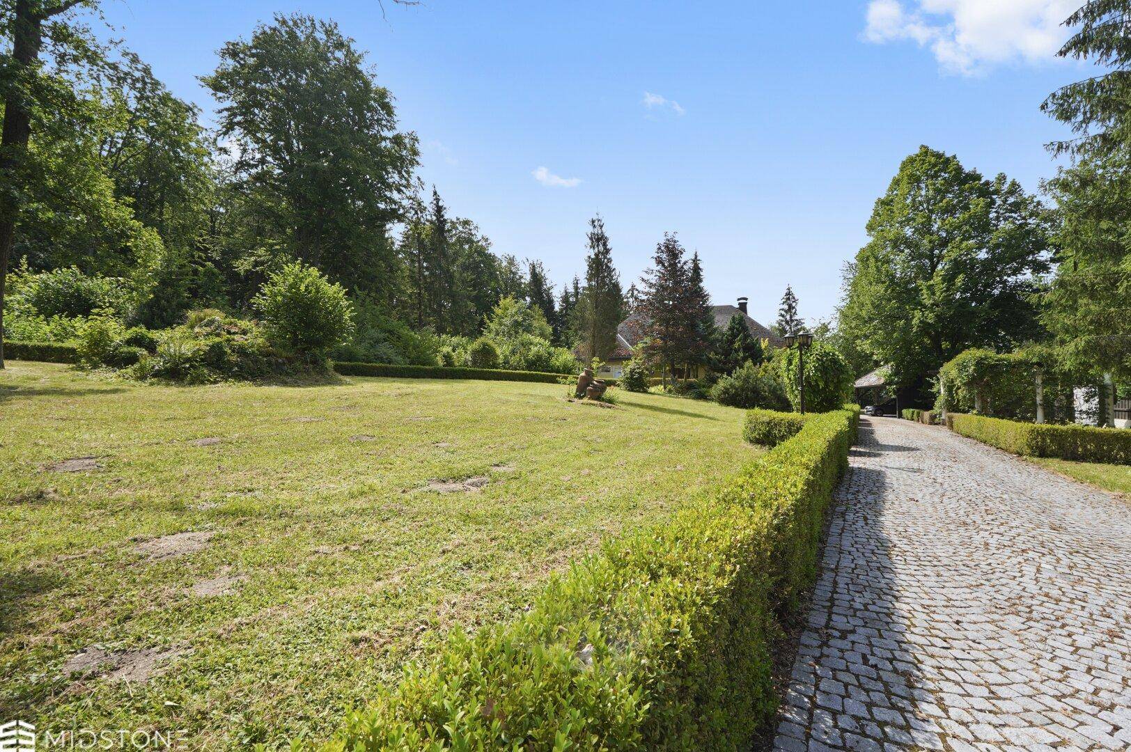 Weitläufiger Gartenbereich mit Rasenfläche, gepflegten Hecken und gepflasterter Auffahrt.