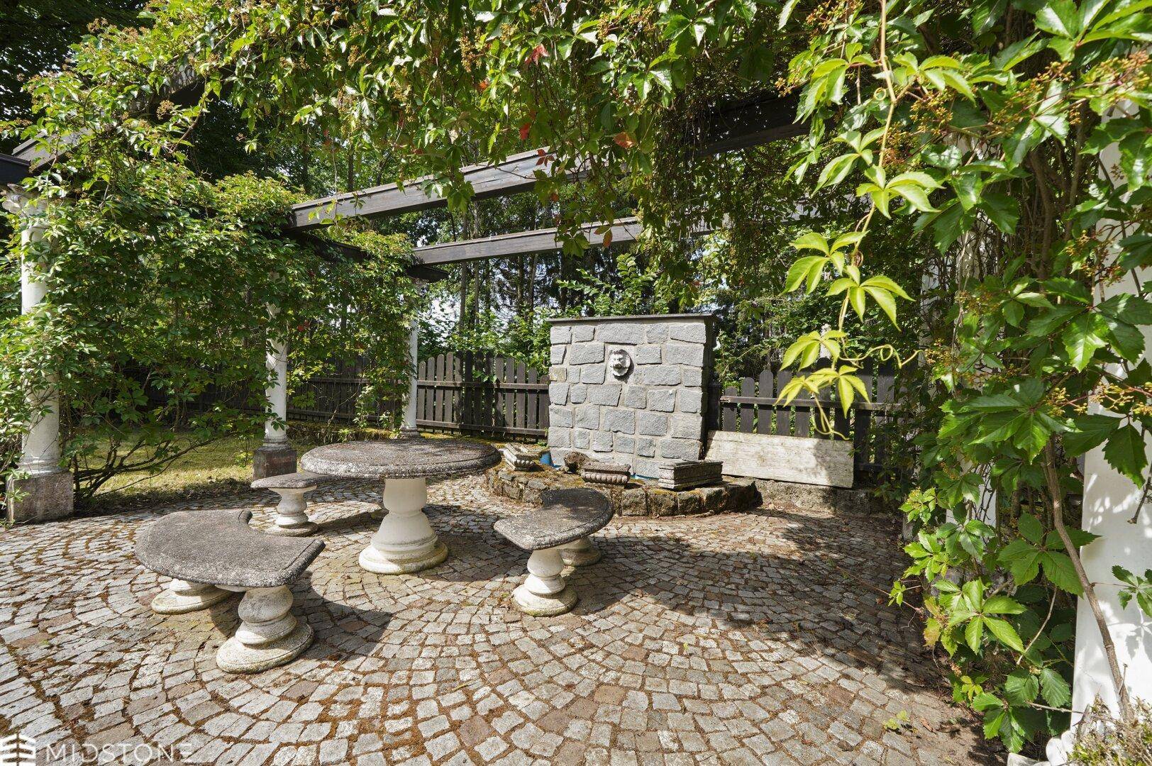 Idyllische Steinterrasse mit Sitzgelegenheiten, Pergola und dekorativem Brunnen im Grünen.