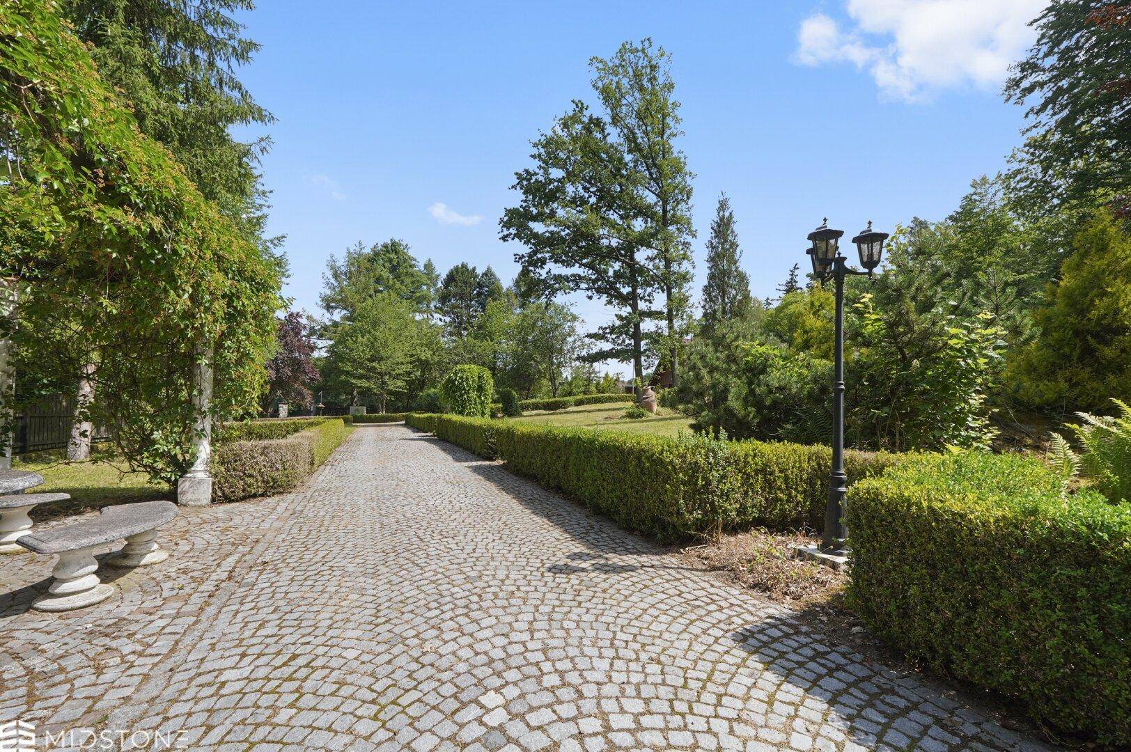 Gepflasterte Auffahrt durch einen gepflegten Garten mit Laternen und Hecken.