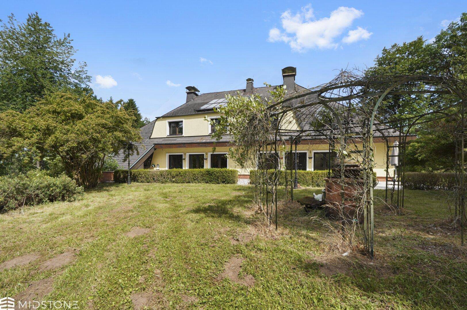 Hinterer Gartenbereich des Hauses mit Rasenfläche, einem Metallbogen und umgebenden Bäumen.