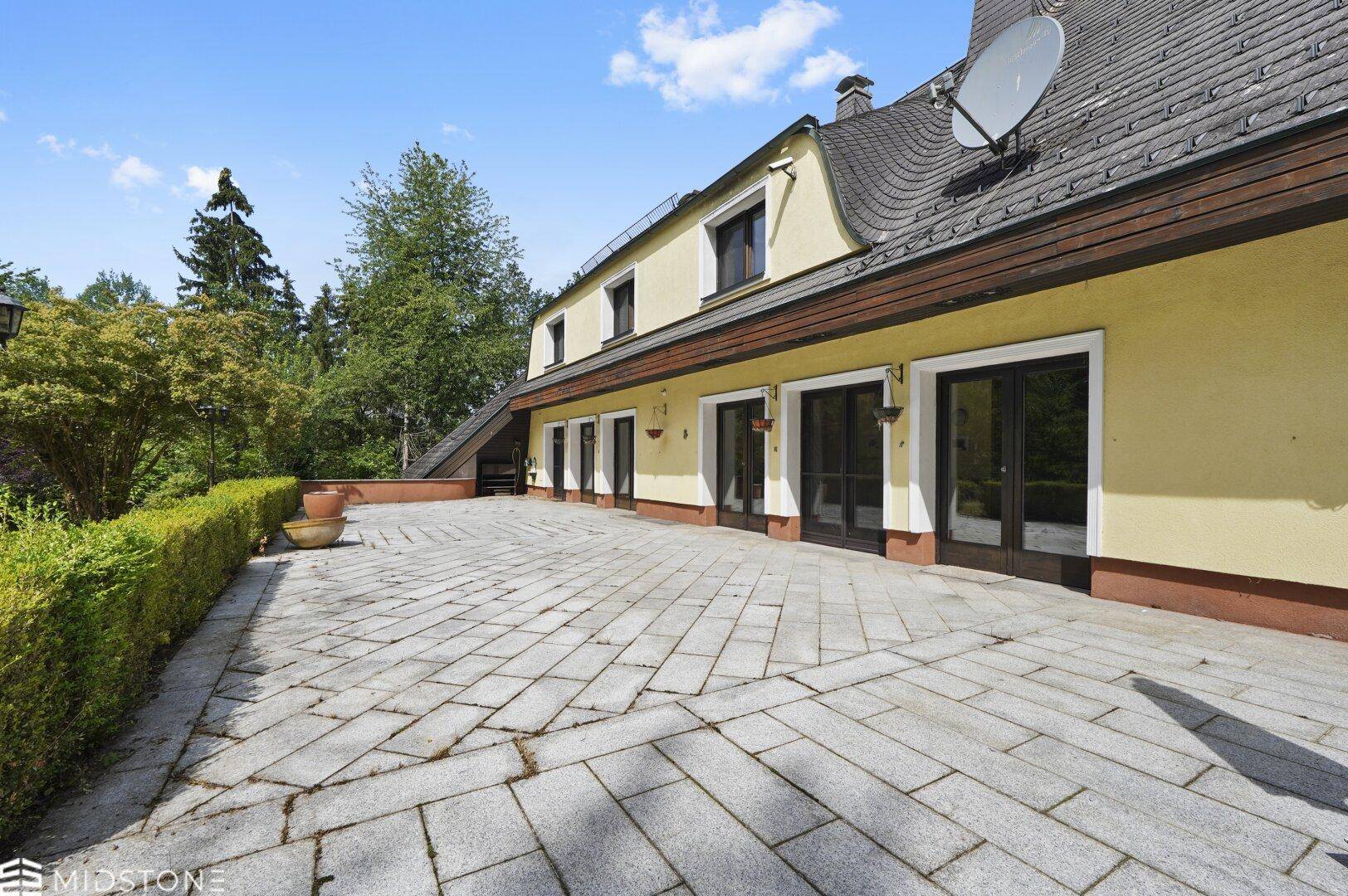 Große gepflasterte Terrasse am Haus mit Zugang zu mehreren Türen und Blick ins Grüne.