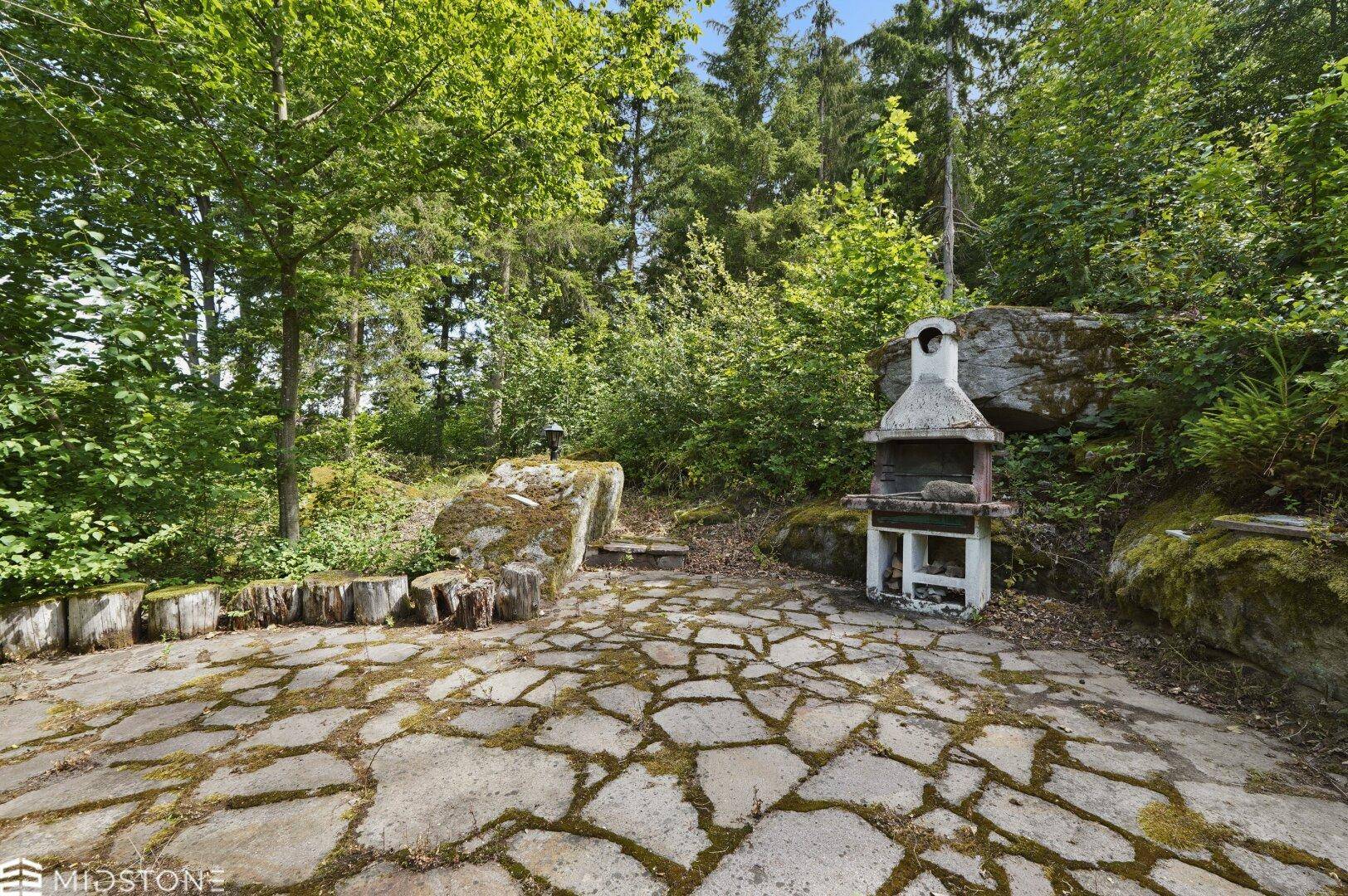 Rustikaler Grillplatz im Garten mit Steinboden und umgebenden Felsen und Bäumen.