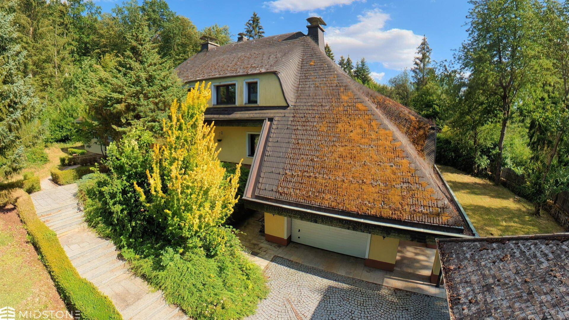 Luftaufnahme des Hauses mit gepflasterter Auffahrt, Garage und umgebendem Wald.