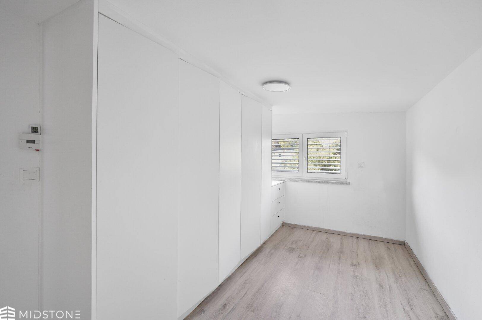 Helles Schlafzimmer mit Einbauschränken, Laminatboden und Fenster mit Jalousien.