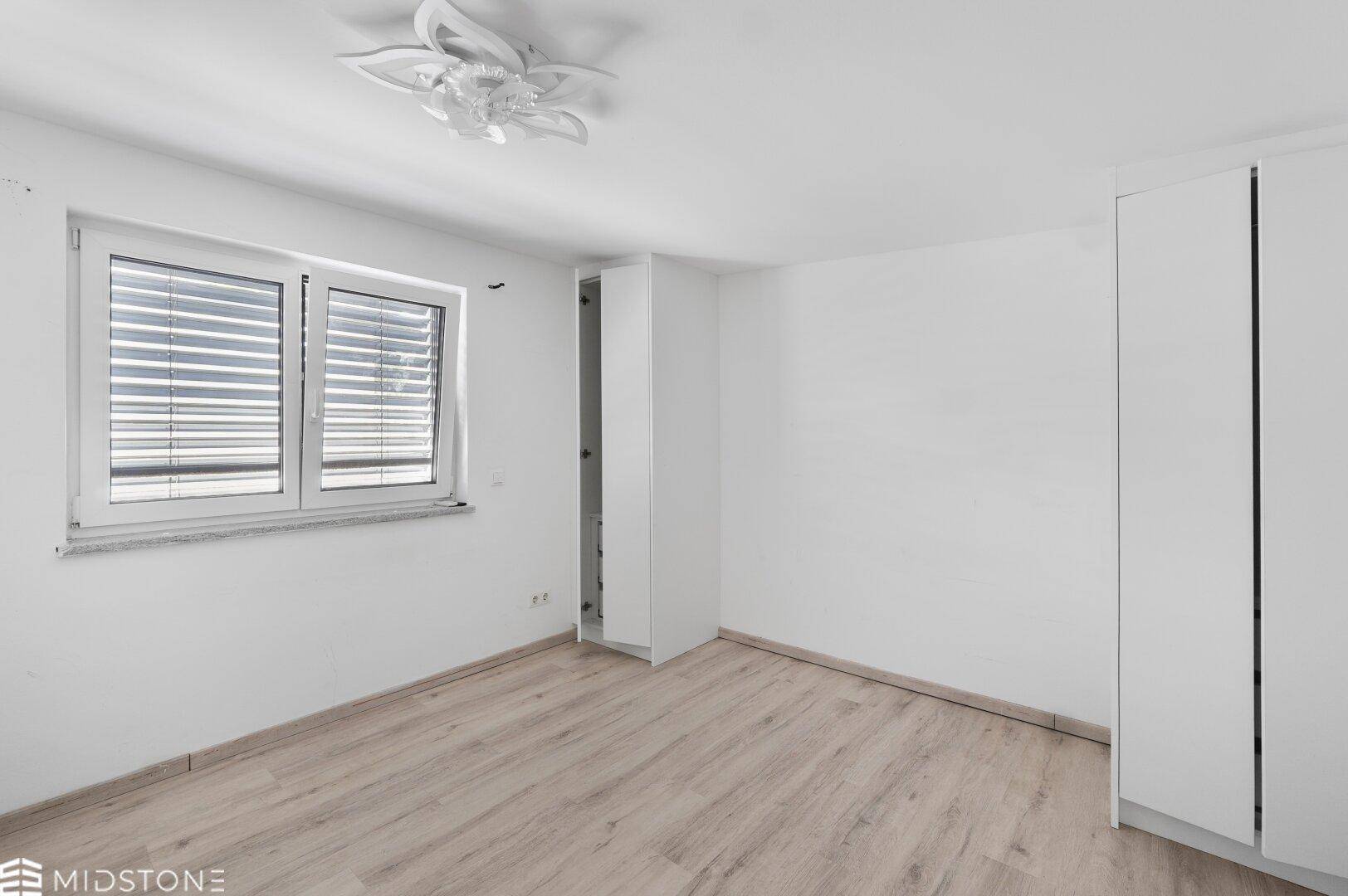 Schlafzimmer mit hellem Laminatboden, Fenster und Deckenleuchte.