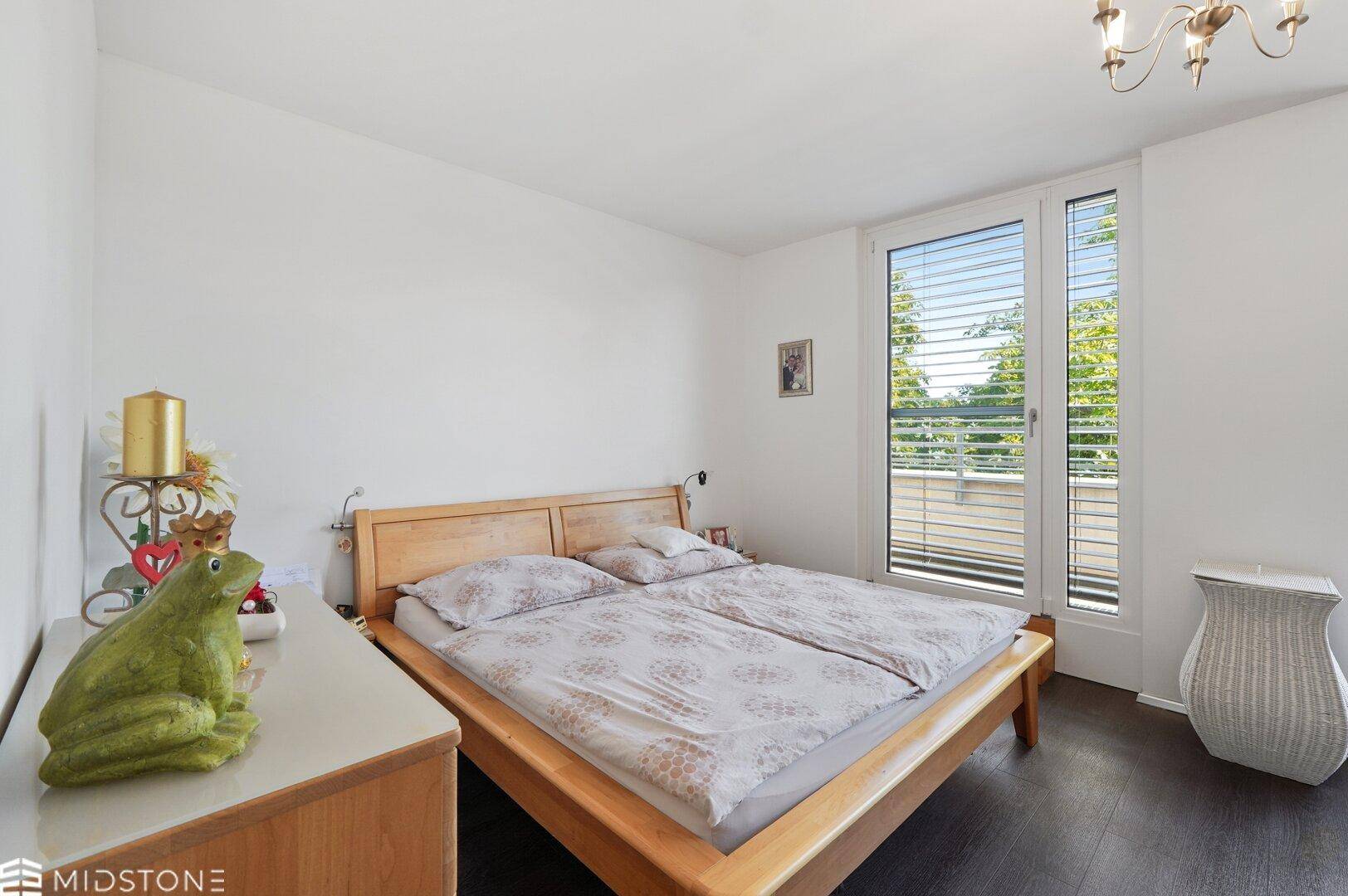 Gemütliches Schlafzimmer mit Holzbett, dunklem Boden und großen Fenstern mit Jalousien.