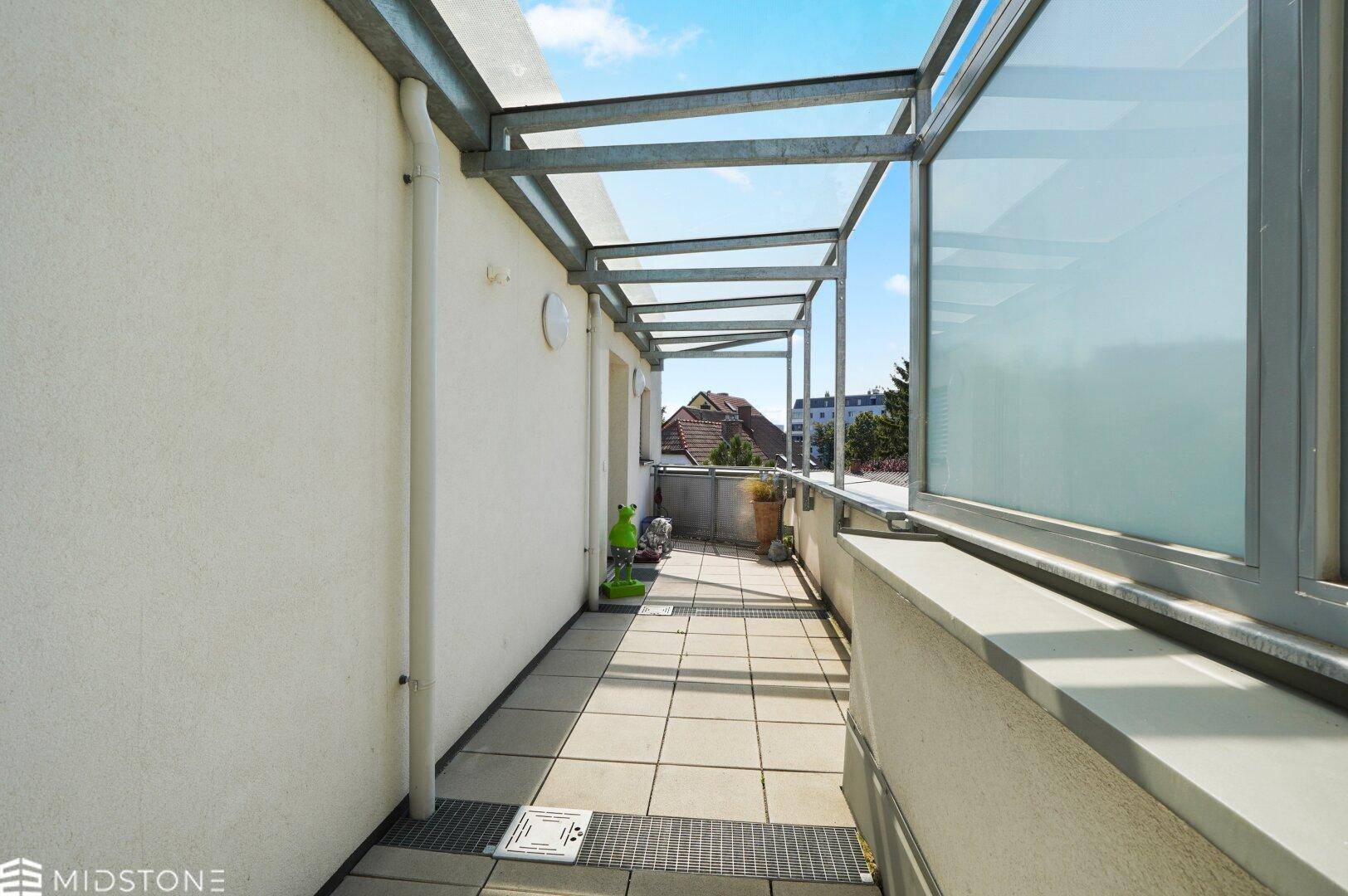 Langer überdachter Balkon mit Fliesenboden und Metallgeländer.