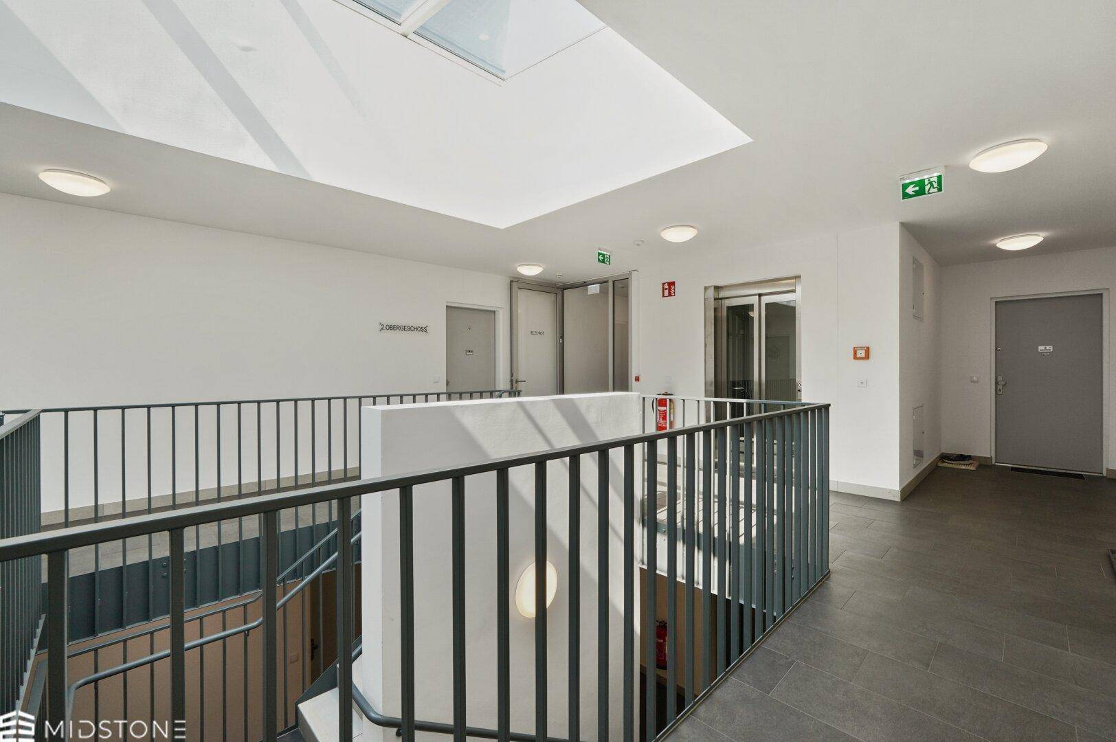 Heller Treppenhausbereich mit Oberlicht, Geländer und Zugang zum Aufzug.