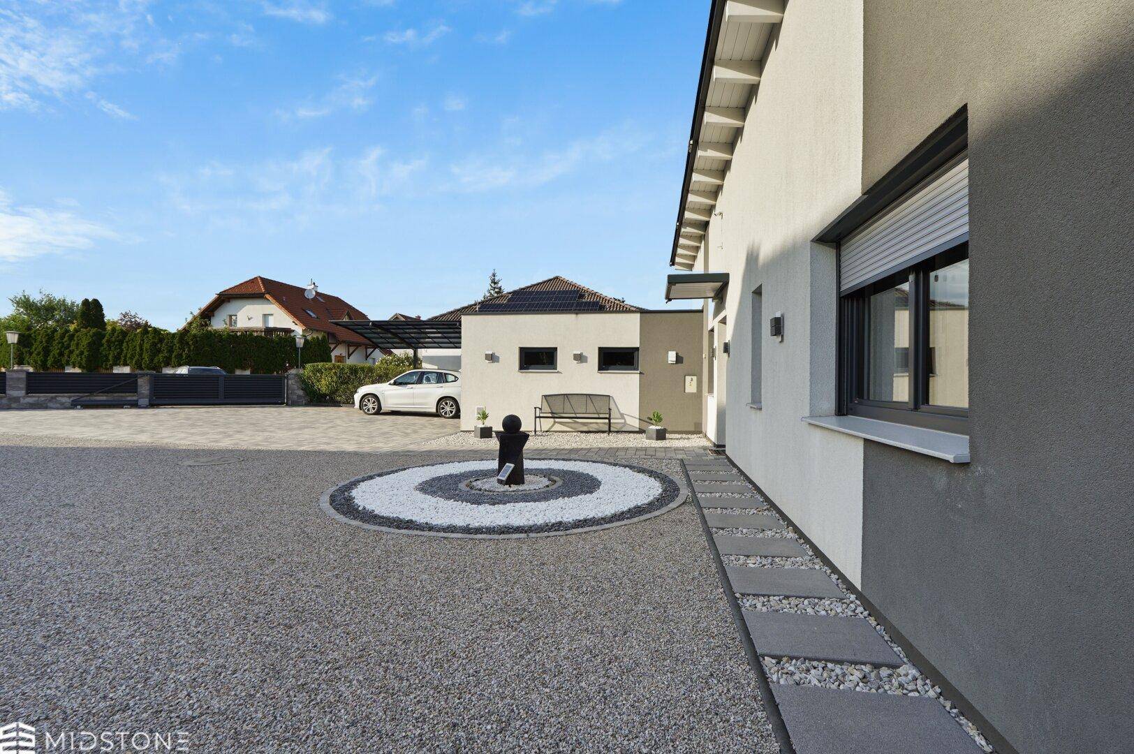 Seitenansicht des modernen Hauses mit Kiesauffahrt, dekorativem Steinkreis und einer Sitzbank im Vorgartenbereich.
