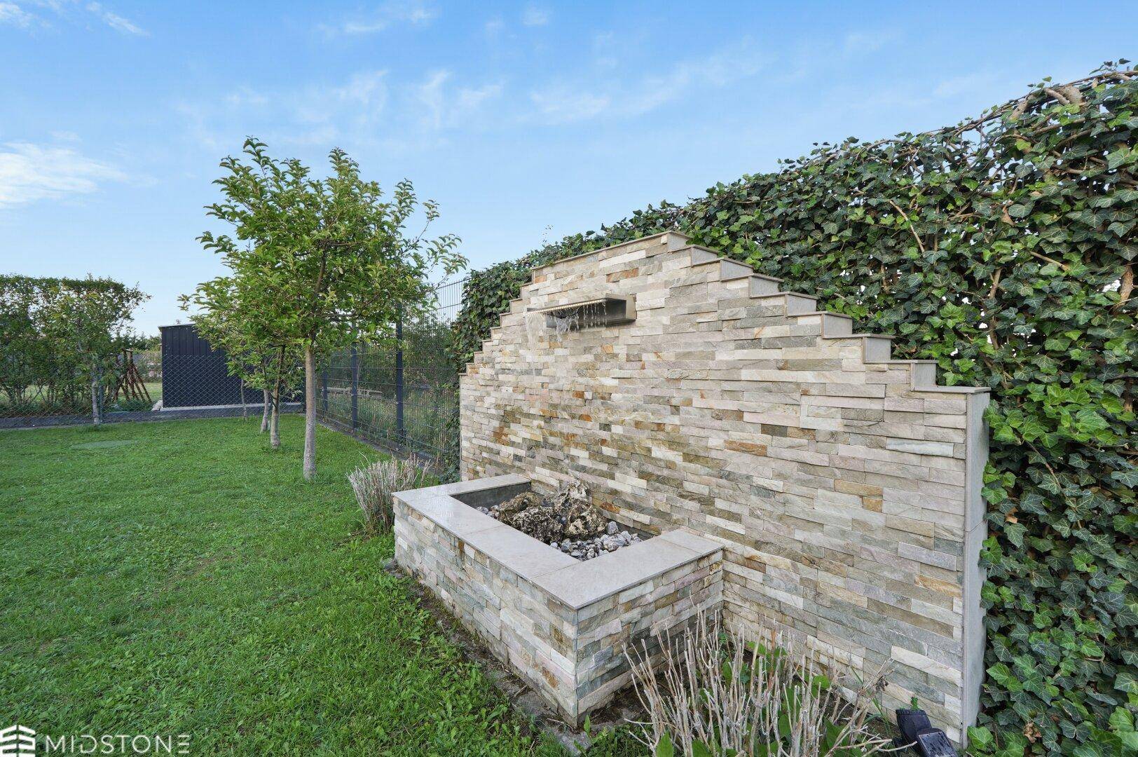 Gartenbereich mit einer dekorativen Steinmauer, integrierter Feuerstelle oder Grillplatz und viel Grün.