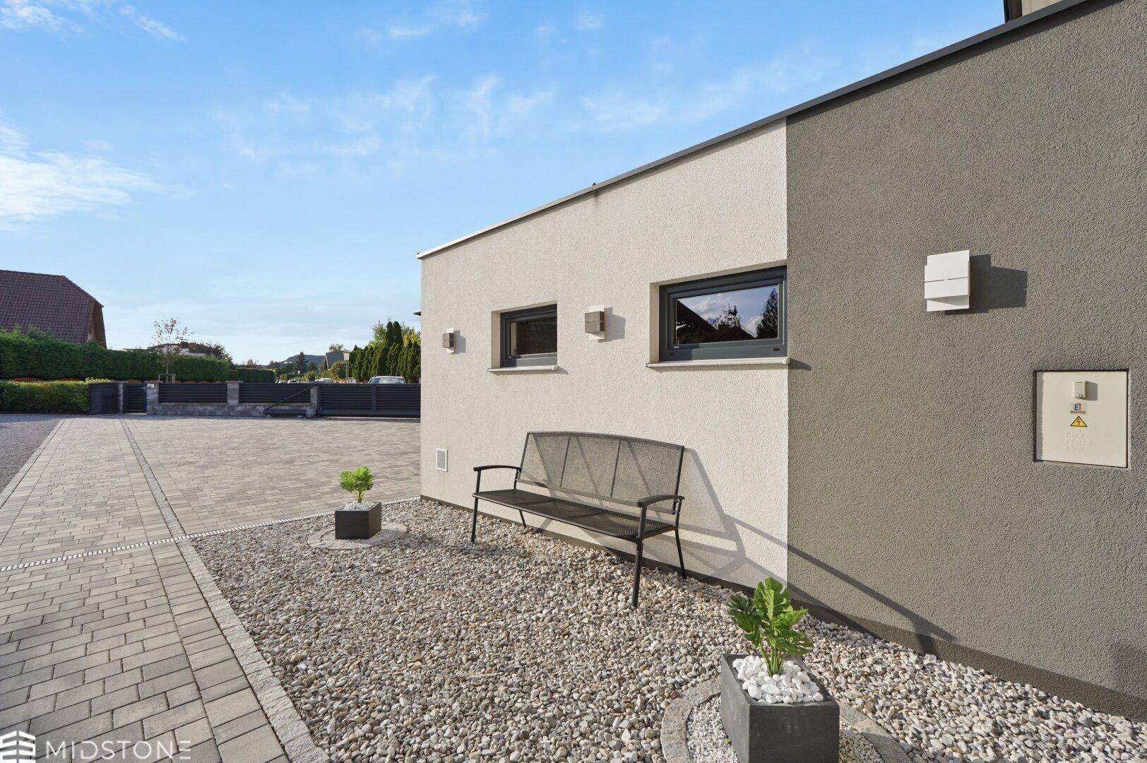Seitenansicht des Hauses mit Kiesweg, einer modernen Sitzbank und kleinen Pflanzen in Töpfen.