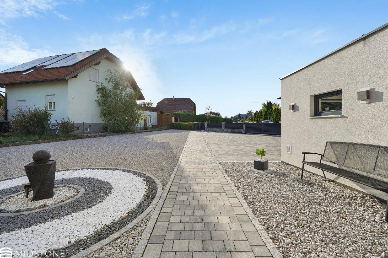 Weitläufige Kiesauffahrt und Vorgarten mit Blick auf ein Nachbarhaus unter strahlendem Sonnenschein.