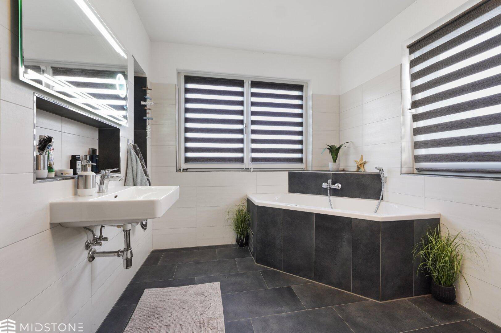 Geräumiges Badezimmer mit Eckbadewanne, großem Spiegel und dunklem Fliesenboden.